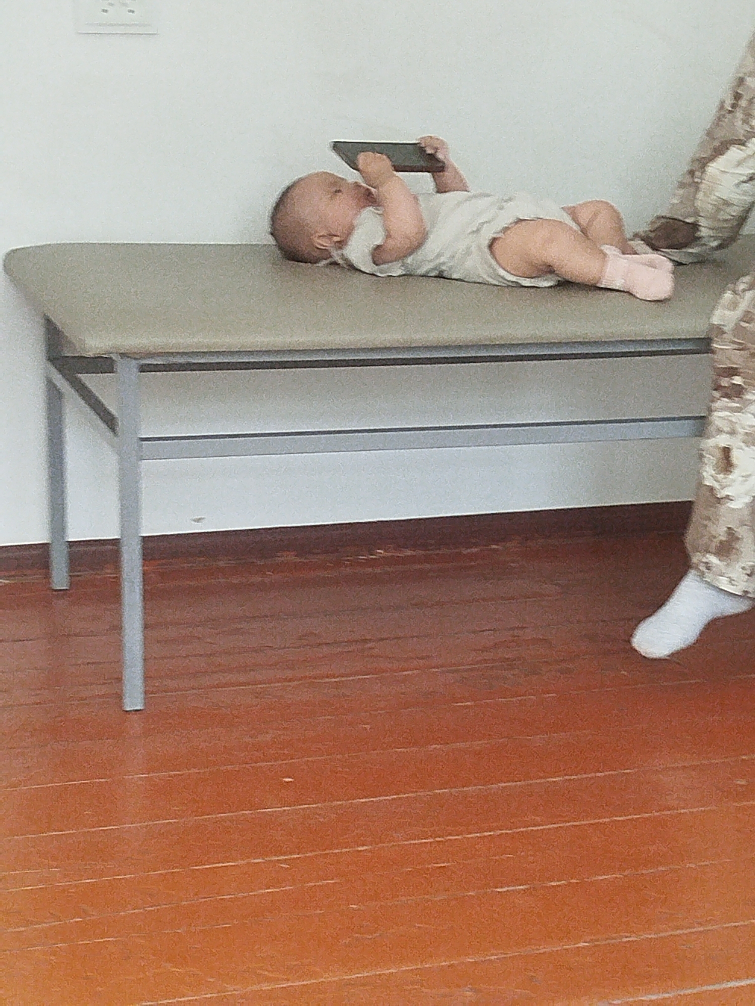 A baby lies on a padded examination table, engrossed in a dark rectangular object, possibly a phone, held in its hands. The baby is wearing a light-colored outfit and pink socks. To the right of the table, a person's legs are visible, clad in camouflage pants and white socks. The examination table is gray with a metal frame. The floor is made of polished reddish-brown wood planks. The background consists of a white wall with an electrical outlet. The scene appears to be an indoor medical setting, potentially in Orozbekov, Kyrgyzstan.