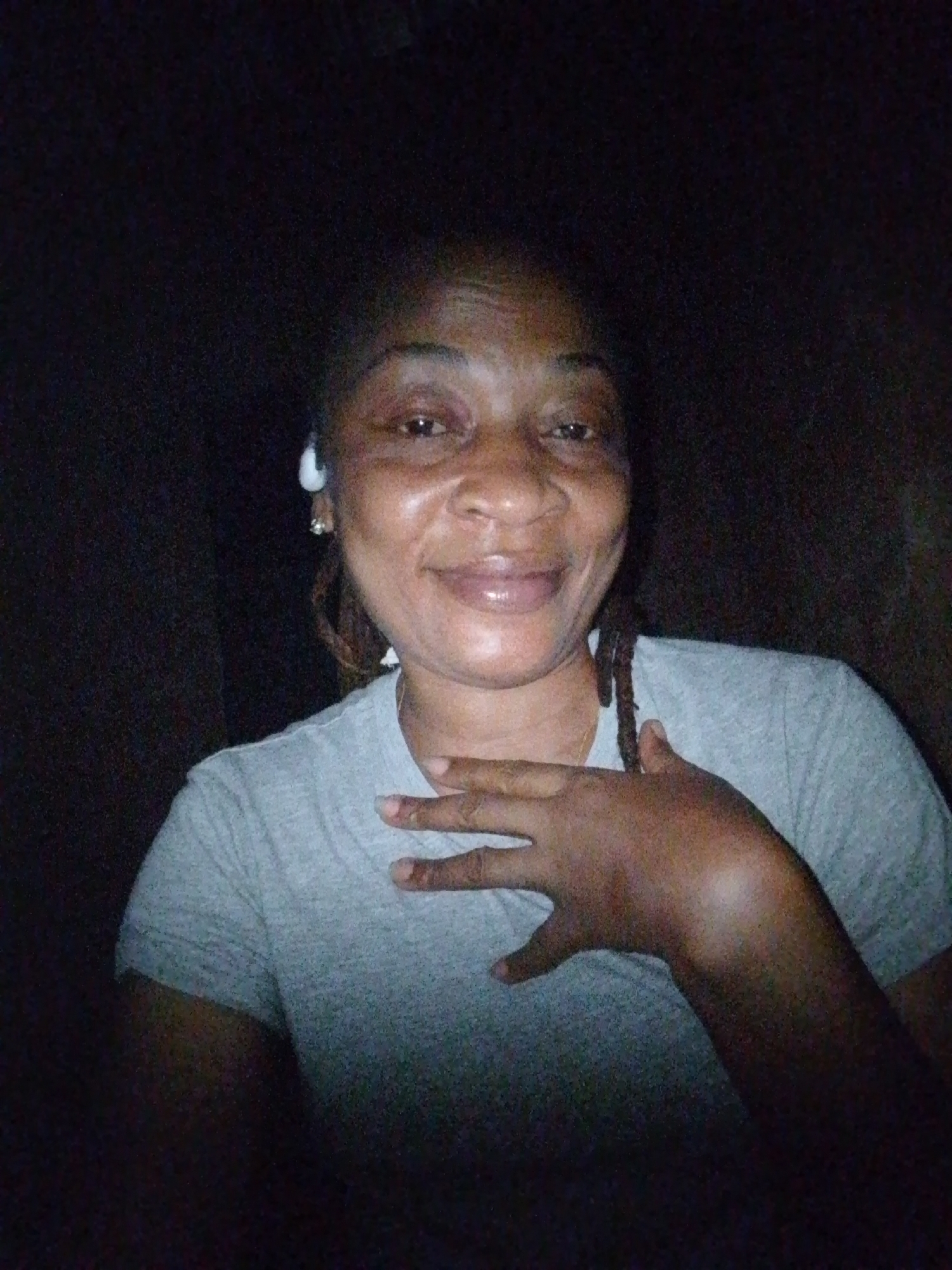 A medium shot captures a dark-skinned woman from the chest up, indoors, with her right hand on her chest and left hand at her side. She is wearing a grey t-shirt and has dreadlocks. White earbuds are visible in her ears. The background is dark and out of focus, with no discernible environmental details. The lighting is dim, suggesting a low-light environment, possibly at night. The woman is looking directly at the camera with a slight smile.