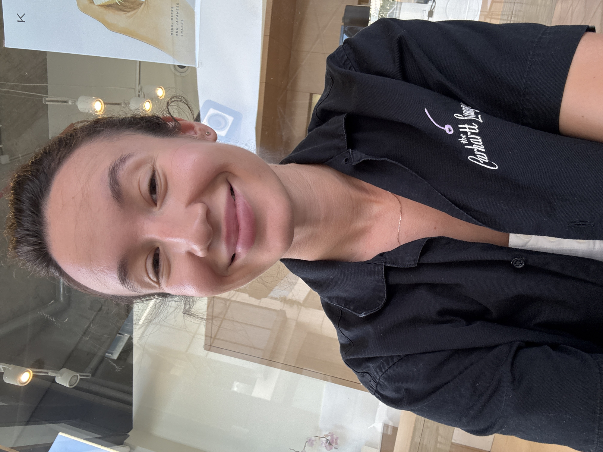 A close-up, selfie-style photograph shows a young woman with her hair pulled back and a wide smile, looking directly at the camera. She is wearing a black button-up shirt with an embroidered logo on the left breast that reads "the Carhartt Lounge" with a cherry icon above the "i" in "Lounge". A delicate gold chain is visible around her neck. The background appears to be an indoor setting with reflective surfaces, possibly a cafe or lounge, with ambient lighting from ceiling fixtures. A poster is partially visible in the upper right corner with text that includes "K" and "WINE, BEERS AND JAPANESE SNACKS". The overall impression is casual and candid. The photographer is identified as being in Tel-Aviv, Israel.