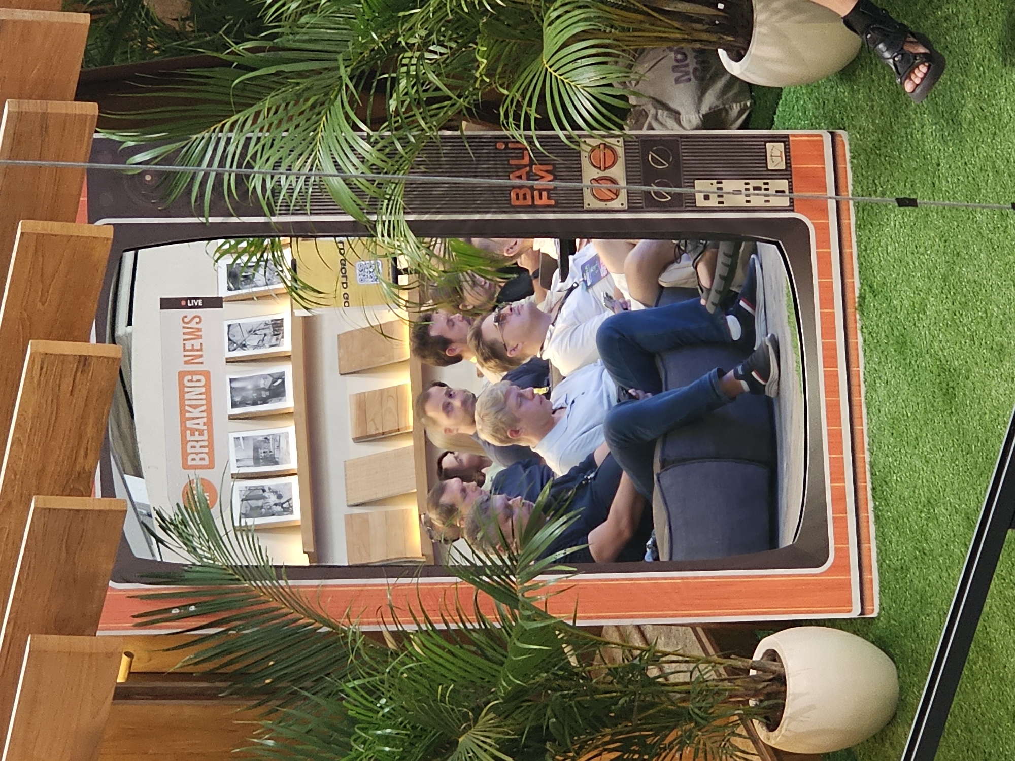 The image shows a group of people sitting on a dark blue sofa, reflected in a mirror in the shape of an old-fashioned TV. The TV frame is orange with a black top, which reads "BALI FM". The reflection shows a "BREAKING NEWS" banner and a series of black-and-white photos on the wall behind the people. The environment includes indoor plants, a wooden structure, and a patch of artificial grass. The image is taken in North Kuta, Indonesia.