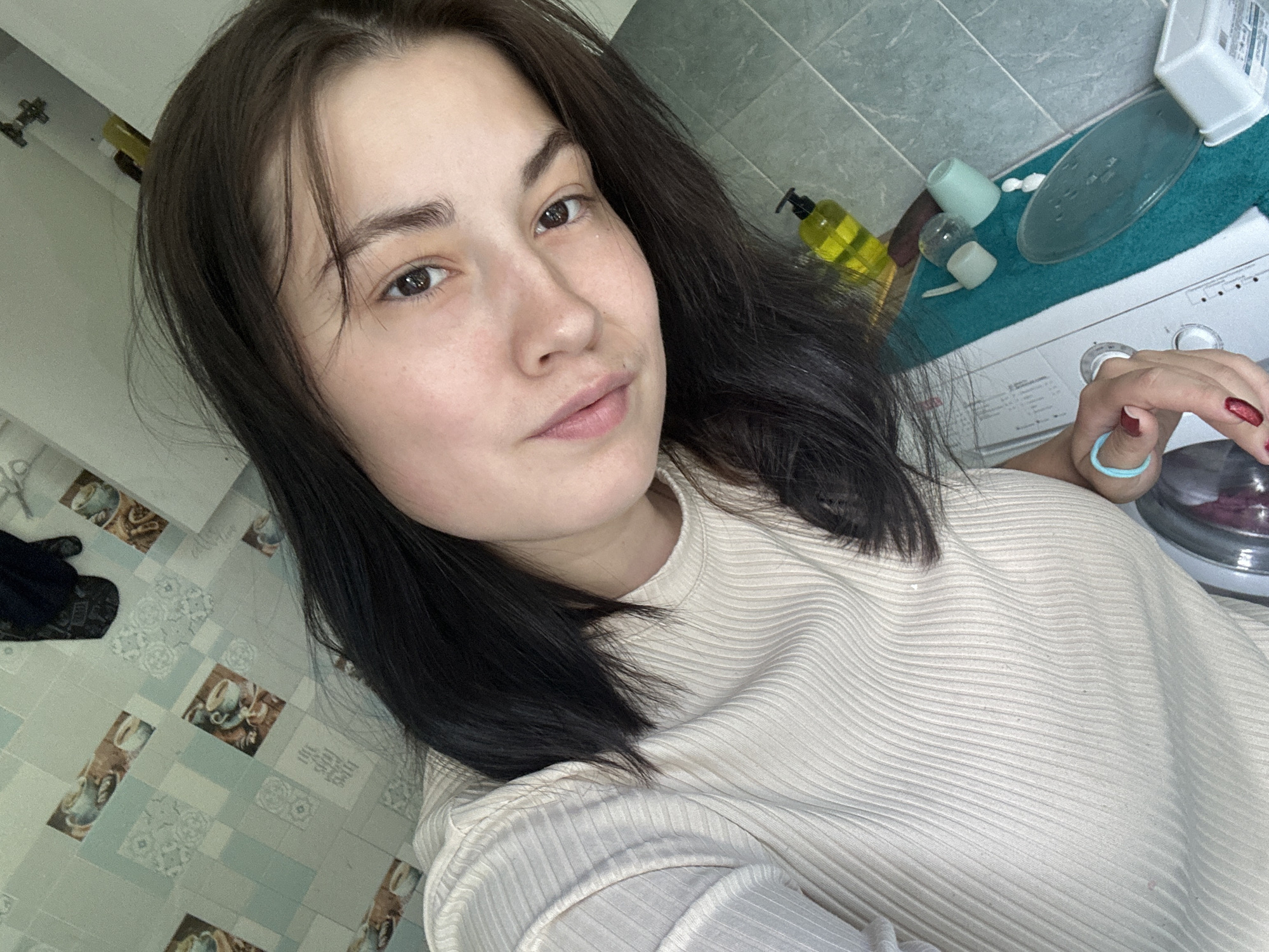 The image shows a young woman with dark hair, wearing a cream-colored ribbed shirt, taking a selfie in what appears to be a bathroom. The background includes tiled walls with a coffee cup motif, a yellow soap dispenser, a washing machine, and various other bathroom items. The overall lighting is bright and natural, suggesting the photo was taken during the daytime. There are no visible indicators suggesting the image was taken in Chaikovsky, Russia, or any specific city.