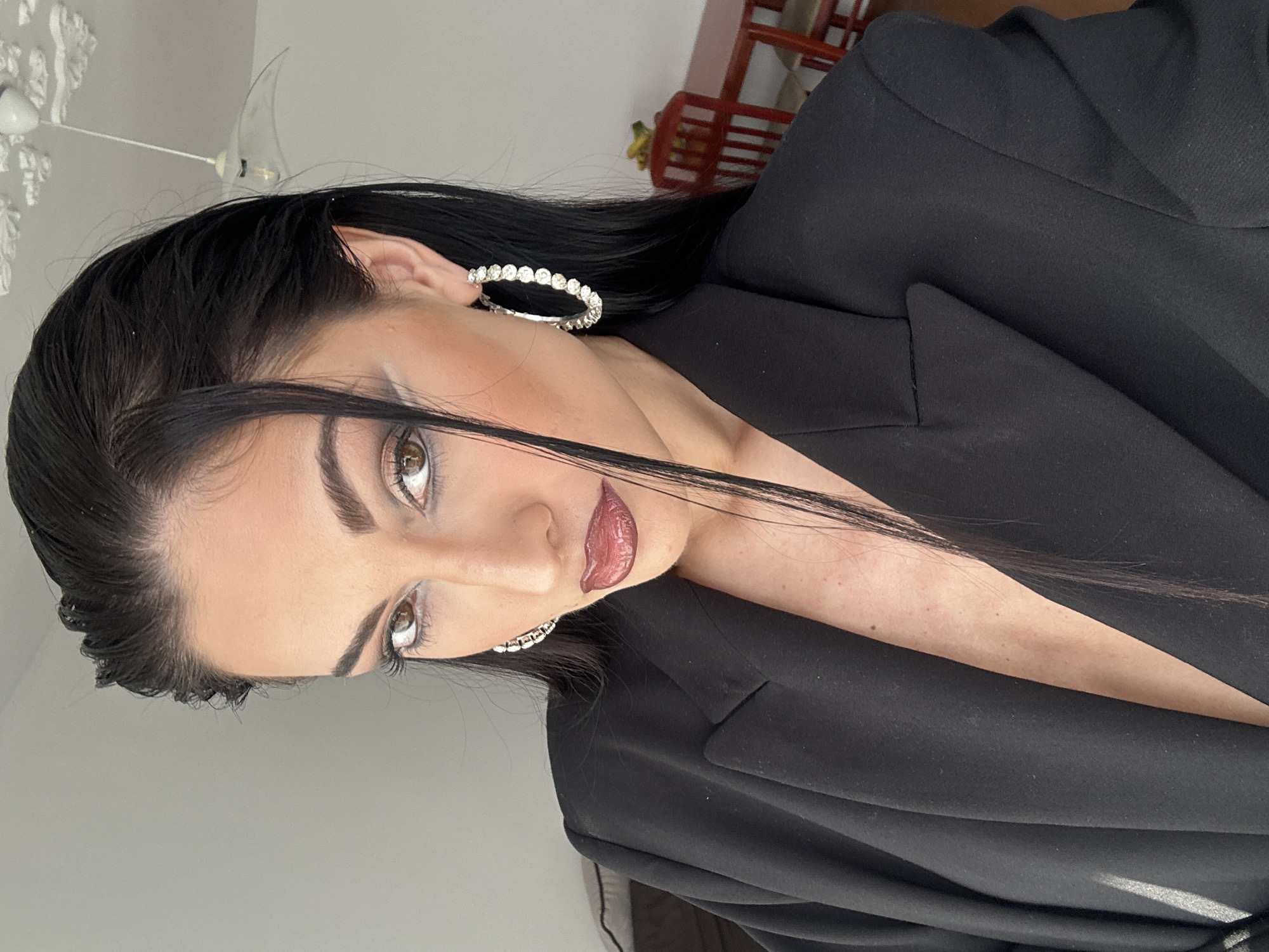 The media file displays a close-up portrait of a female, rotated 90 degrees clockwise. She has long, dark hair, styled to the side, and is wearing a dark, likely black, blazer or jacket. Her head is tilted, and she gazes downwards and to the left with a neutral yet focused expression. She wears striking silver-toned hoop earrings, each embellished with a series of small, sparkling stones. Her facial makeup is prominent, featuring defined eyebrows, dark eyeshadow, winged eyeliner, and glossy, reddish-brown lipstick.
The lighting appears soft and even, highlighting the sheen on her lips and the reflective qualities of her earrings. In the background, a light-colored ceiling with a white, decorative molding or a partial light fixture is visible towards the top-left of the rotated image. A reddish-brown wooden structure, possibly a chair back or shelving unit, can be seen in the top-right. The setting suggests an indoor environment with plain, light-colored walls. The subject is actively posing for the photograph. No visible text is present.