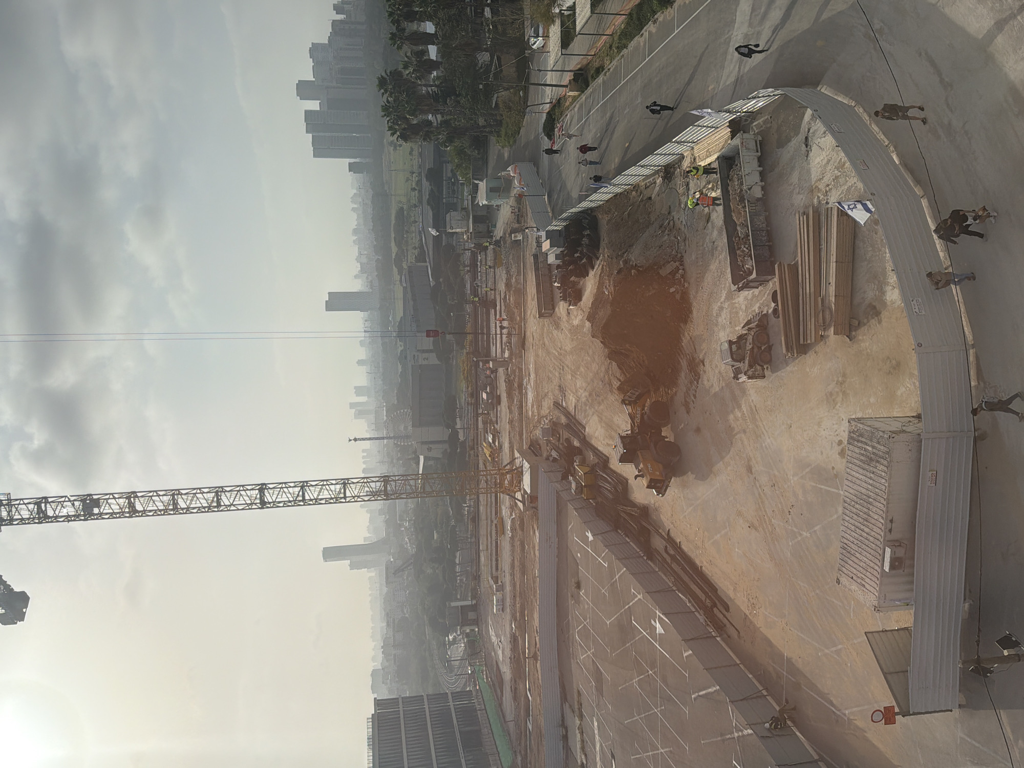 The image depicts a construction site in Tel-Aviv, Israel. Visible is a large crane extending over the construction area. In the background, there are a number of high-rise buildings, likely part of the Tel-Aviv skyline. In the construction site itself there are various construction materials, and people are moving around and performing tasks. A fence is visible. An Israeli flag is present.