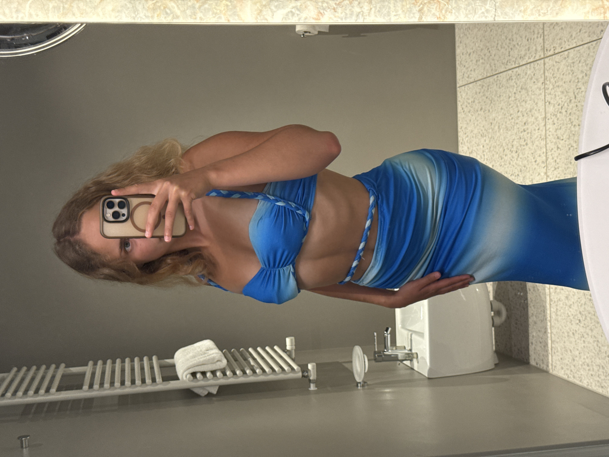 In a well-lit bathroom, a young woman with blonde wavy hair poses for a mirror selfie. She is wearing a blue and white ombre two-piece outfit consisting of a ruched strapless top and a long, form-fitting skirt. Her phone, held in her right hand, is angled to capture her reflection. The bathroom features a white wall, a towel rack with a folded white towel, a sink with a faucet, and a tiled floor. The overall aesthetic suggests a modern and clean environment. This scene does not have any apparent connection to the city of Trieste, Italy.