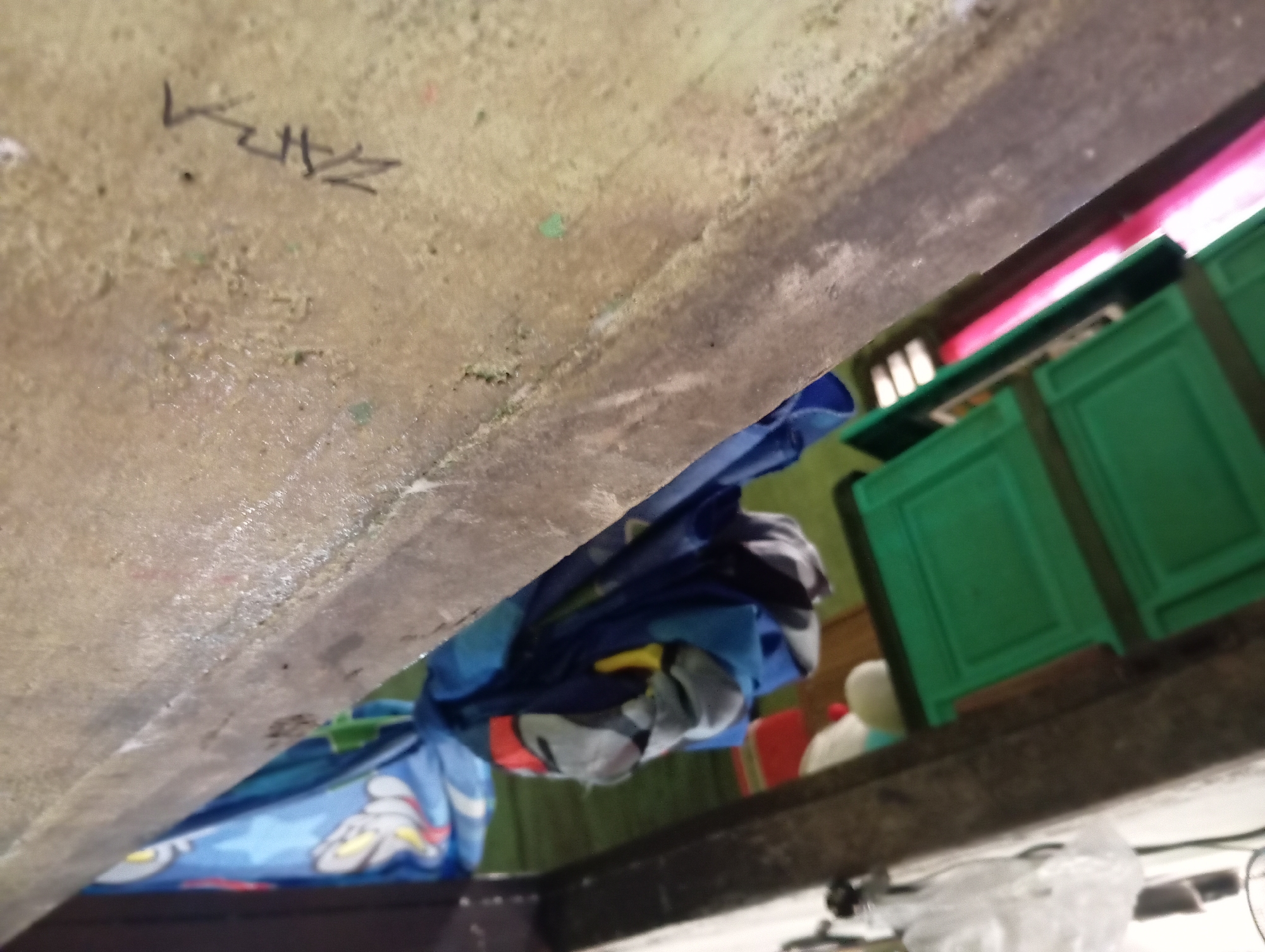 In Rejosari, Indonesia, the image captures an outdoor scene, angled from above, showing a close-up of a weathered concrete ledge. Below the ledge, a blue tarp with a cartoon character print hangs, partially obscuring green plastic storage drawers. A pink object is visible behind the drawers. The concrete ledge is marked with black graffiti and shows signs of wear and staining. The overall impression is of a somewhat cluttered, utilitarian space.