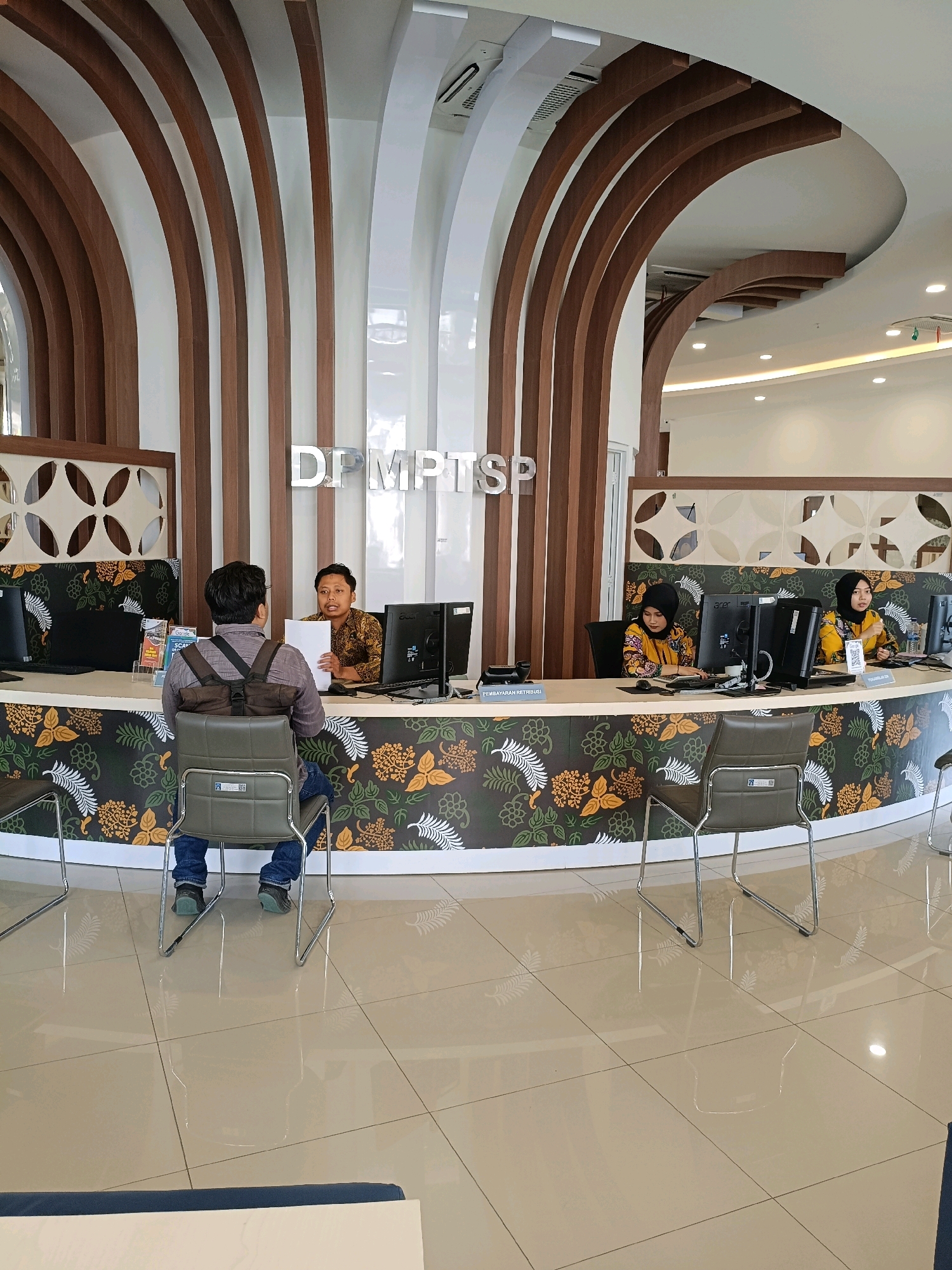 In a government office in Sleman, Indonesia, a male customer sits facing a male employee behind a reception desk, holding a document. The desk is decorated with a floral pattern and computers are visible. Two female employees sit at computers on the other side of the reception desk. The office has a modern design with curved wooden accents and tiled floors.