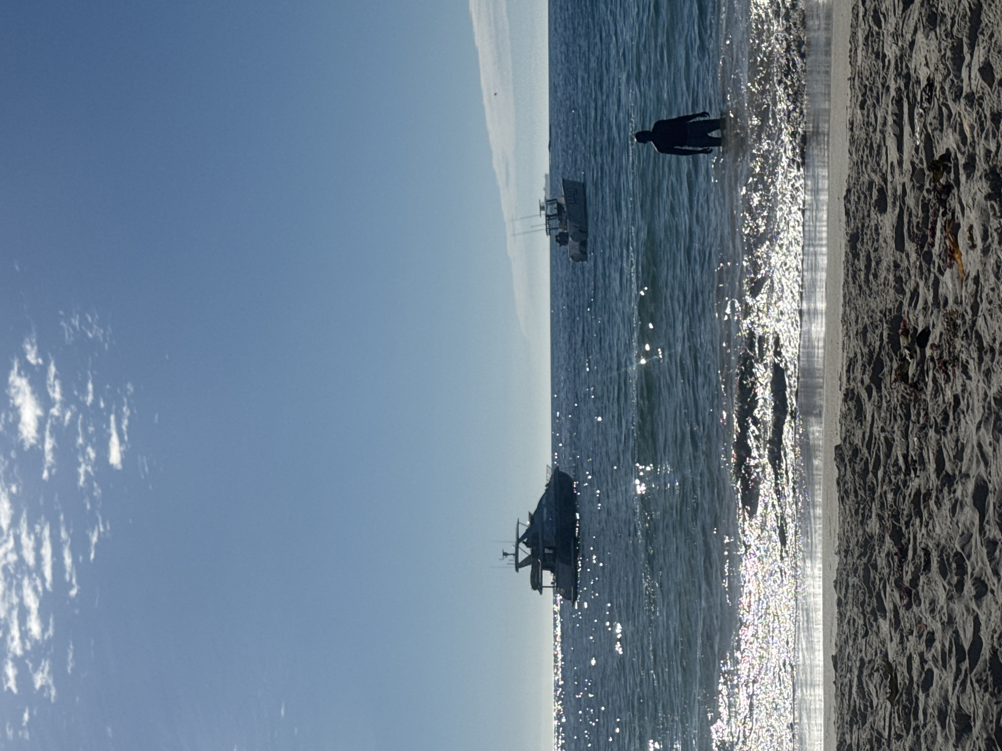 The image displays a bright, sunlit coastal scene in Cape Town, South Africa. The foreground features textured sandy beach with visible ripple patterns and minimal organic debris. The ocean occupies the mid-ground, exhibiting a dark blue coloration with numerous intense, glinting reflections of sunlight across its surface, indicative of strong backlighting. A solitary individual, appearing as a dark silhouette, stands waist-deep in the ocean, facing away from the viewer towards the horizon. This person is wearing dark-colored shorts or swimwear. Two boats are visible in the water: a larger, dark-hulled yacht or cabin cruiser is positioned to the left, and a smaller, white-hulled fishing boat with the name "HALO" on its side and fishing rods attached is situated to the right. Both vessels appear stationary or adrift. On the distant horizon to the right, a large cargo ship is faintly visible. The sky above is clear blue with sparse, thin white cloud formations.