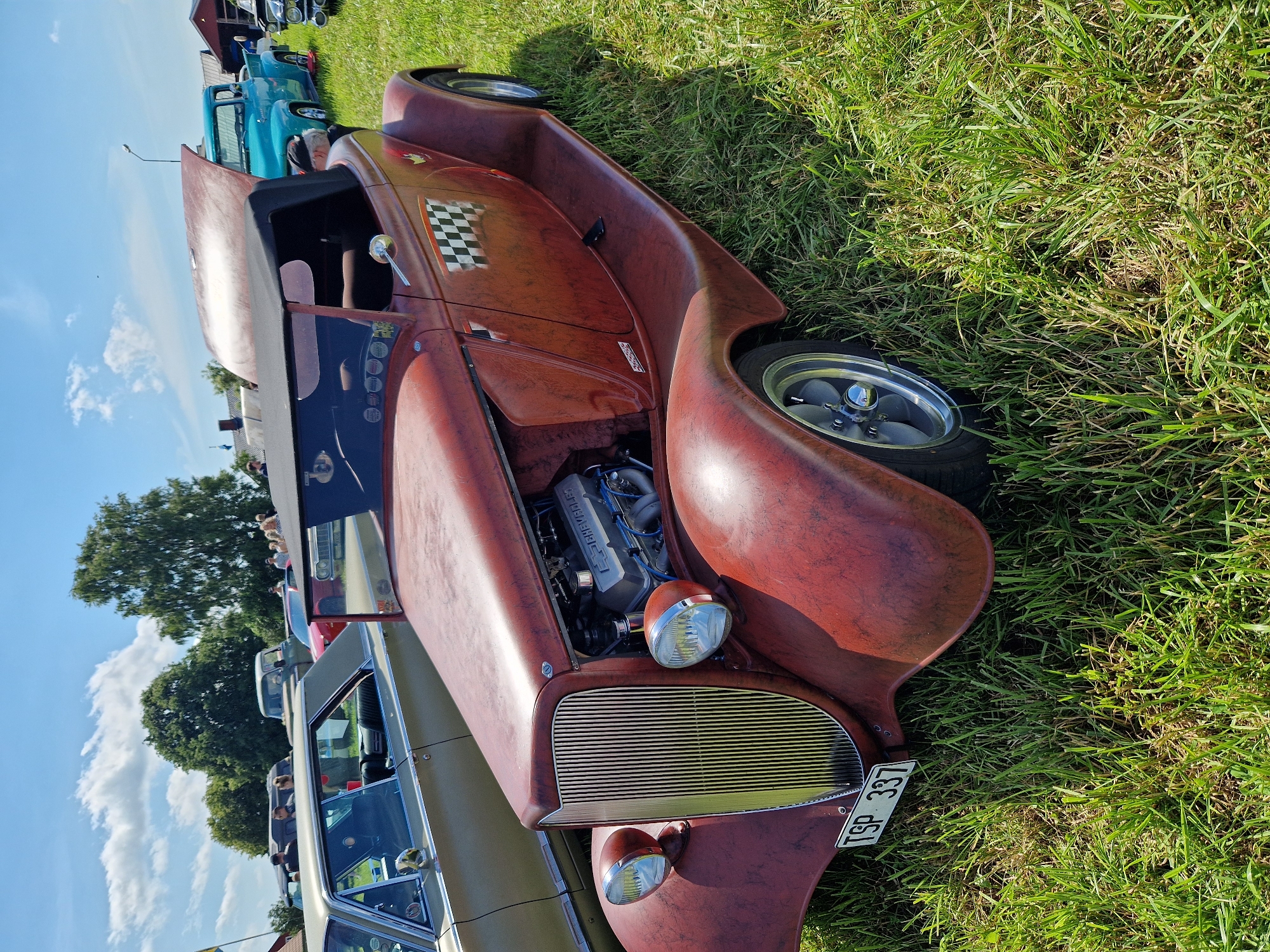 In Edenberga, Sweden, on a grassy field under a partly cloudy sky, a red hot rod convertible is parked. The car features a distressed red paint job, a black convertible top that is up, a prominent chrome grille, and exposed engine parts. A checkered flag decal is visible on the side of the car. The license plate reads "TSP 337". Other vehicles, including a light blue pickup truck and a tan sedan, are present in the background, along with a few people mingling. The environment appears to be an outdoor gathering or car show.