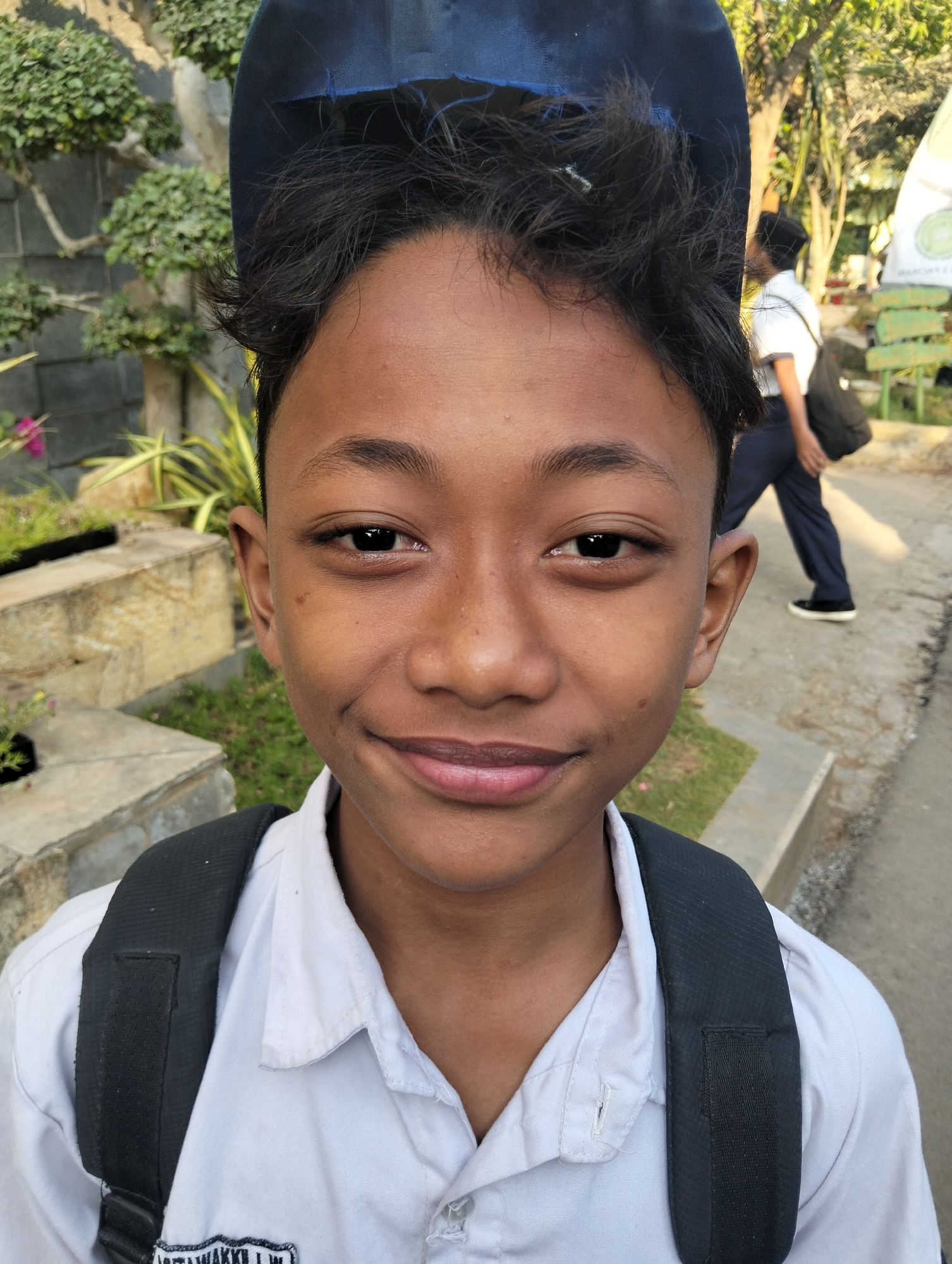 In an outdoor, possibly urban or suburban, setting with visible foliage, trees, and concrete structures, a young male is the central subject of the image. He is looking directly at the camera, with a slight smile. He is wearing a white collared shirt, a black backpack with visible straps, and a dark blue cap worn backwards. His dark hair is slightly disheveled. In the background, another person wearing a white shirt and dark pants is walking away from the camera, carrying a black bag. The environment suggests a daytime scene with natural lighting. The image appears to be taken in Paciran, Indonesia, as indicated by the text visible on a patch on the subject's shirt.