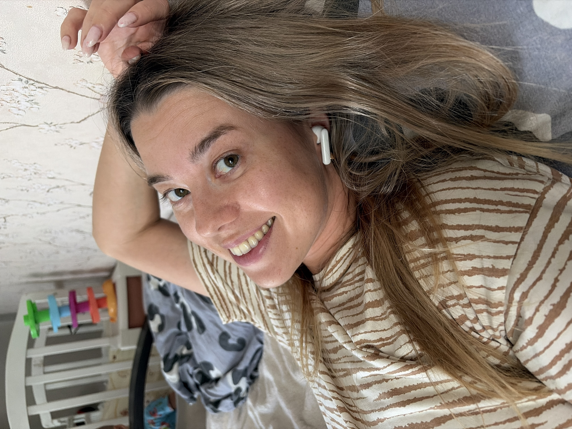 A woman lies on a bed, her head propped up on her arm, with her hand resting on her head. She is looking at the camera with a smile and her eyes are wide open. She is wearing a striped shirt and has earbuds in her ears. The background is a wall with floral wallpaper and a crib with a colorful toy. The scene appears to be indoors, possibly in a bedroom, in Chaikovsky, Russia.