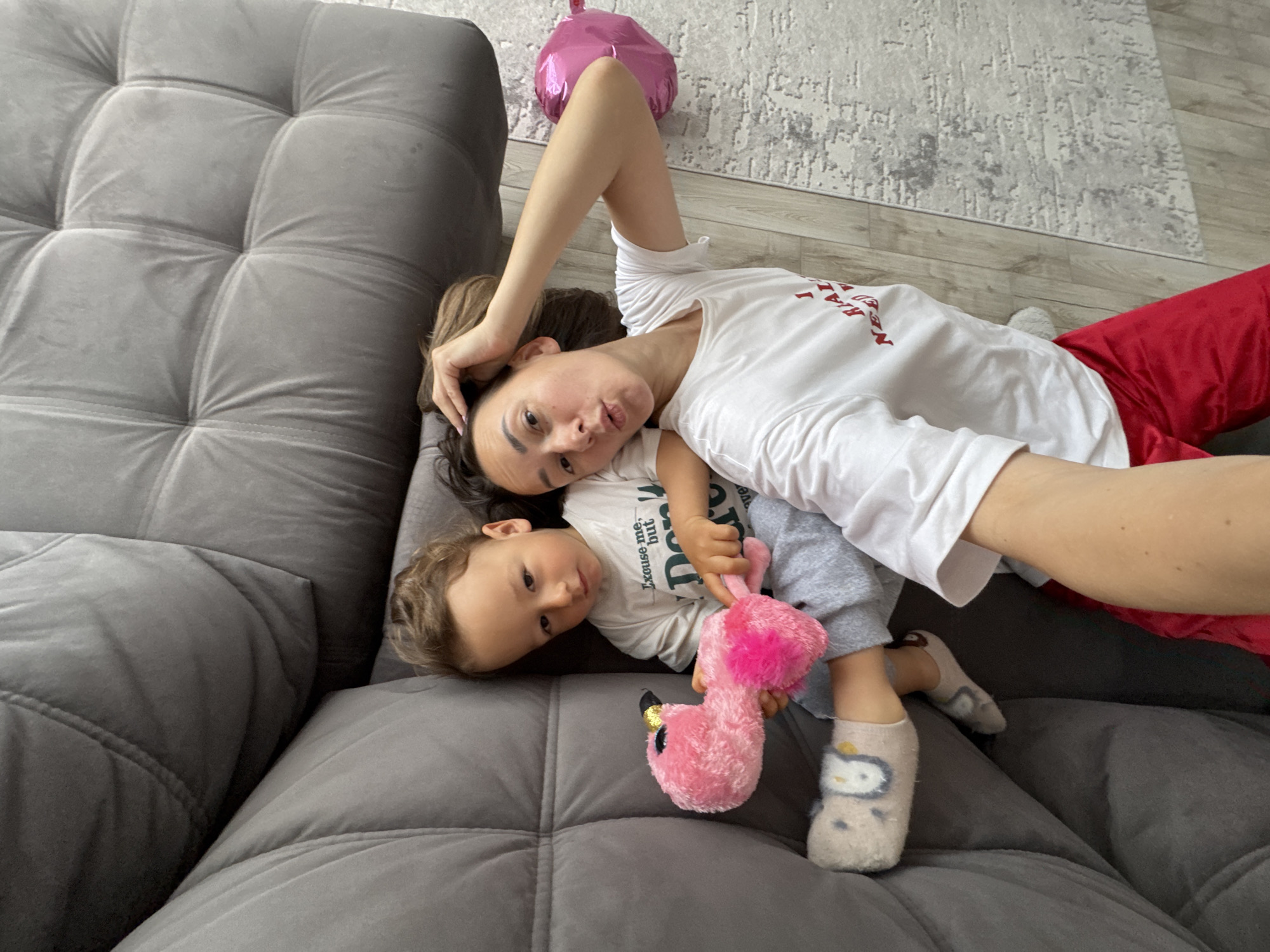 A woman and a young child are lying on a grey, tufted sofa. The woman is wearing a white t-shirt with red writing and red pants. She has her left arm above her head. The child is wearing a white t-shirt with green writing, grey pants, and socks with a cartoon character on them. The child is holding a pink stuffed flamingo. A pink balloon is visible in the background, near a rug on the floor. The location is potentially a home in Sochi, Russia.