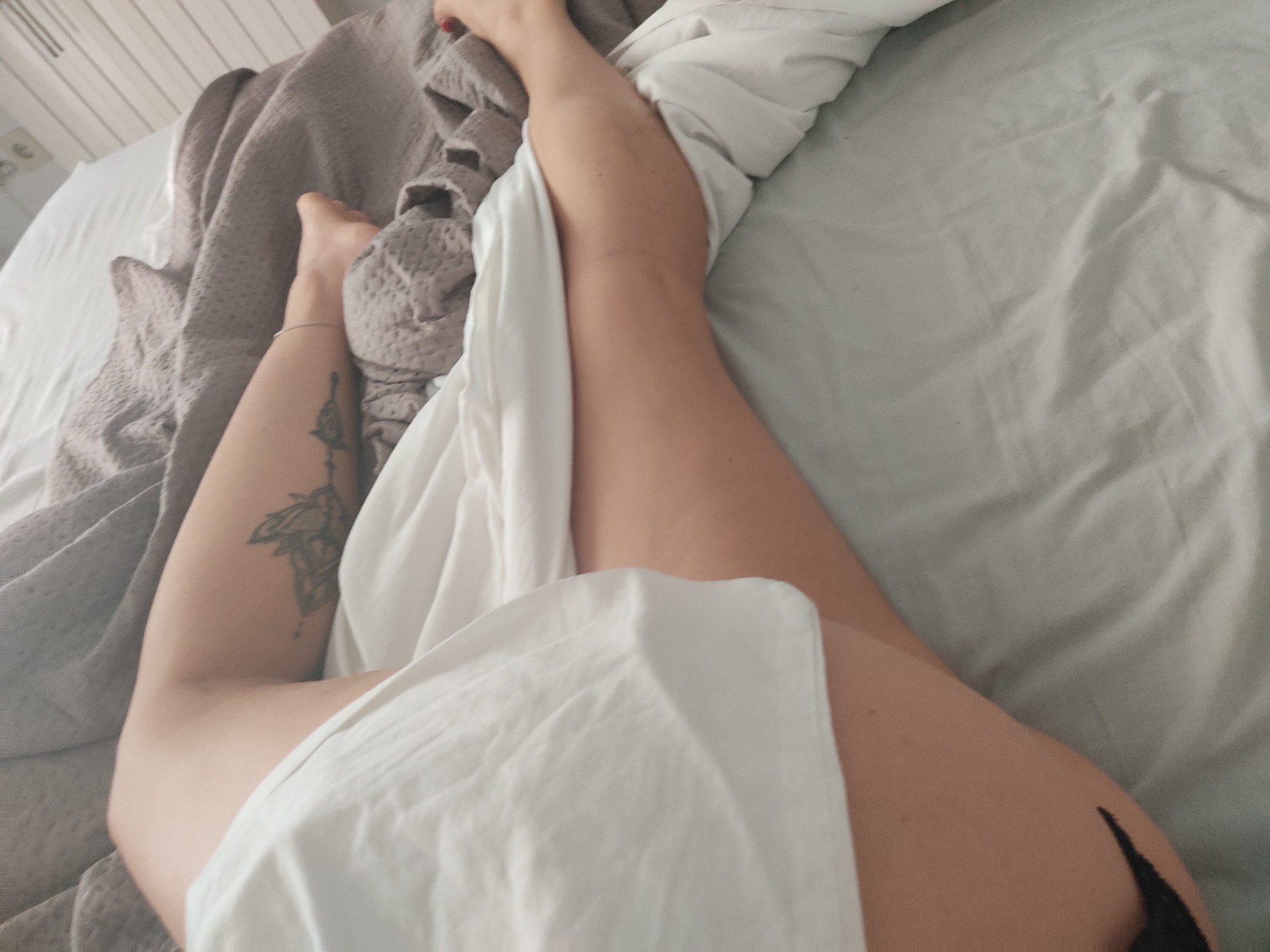 The image shows a person lying in a bed. The person's legs are visible, covered partially by a white sheet. A grey blanket is draped over the bed. A tattoo is visible on the person's left thigh. The scene appears to be indoors, likely a bedroom. There is no information in the image to suggest the location is Blanes, Spain.