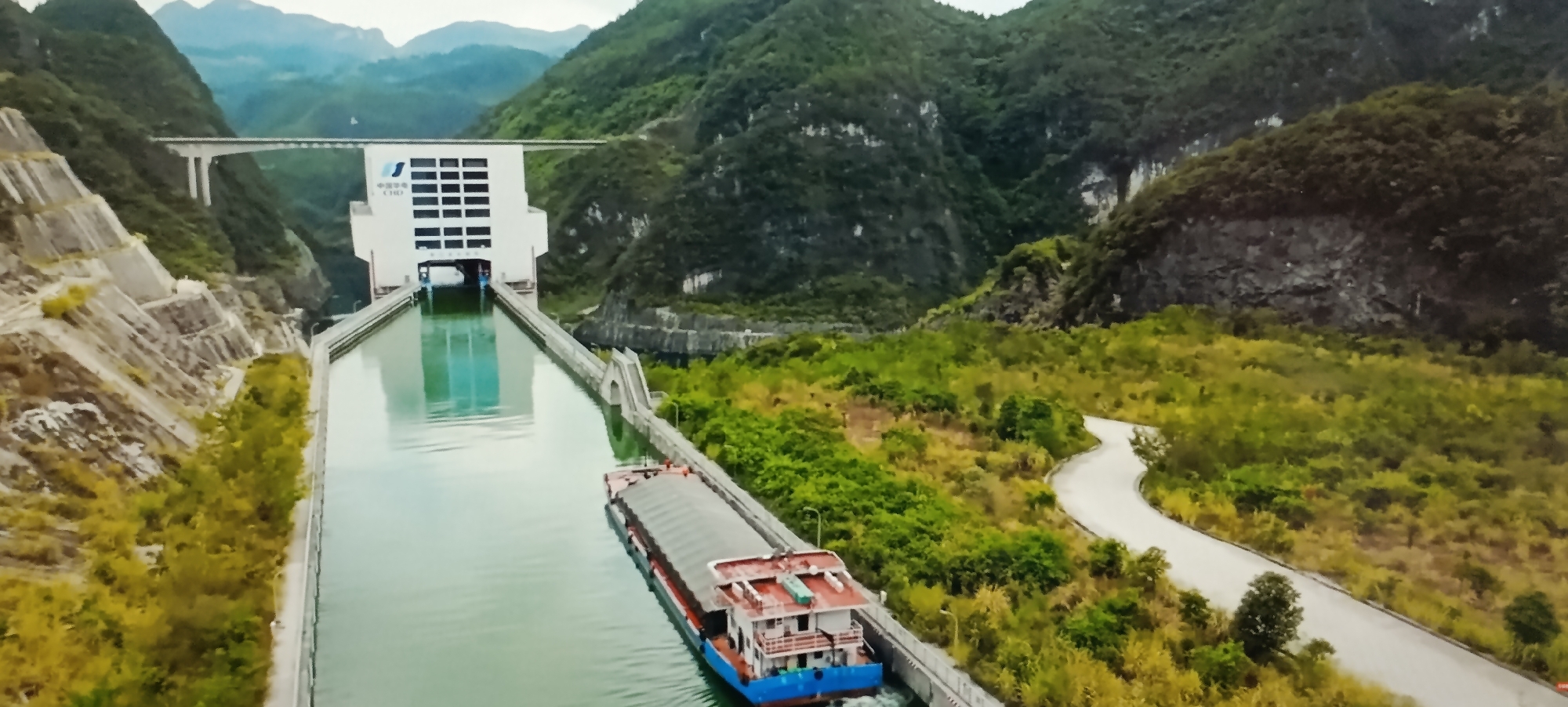 The image shows a view from above of a large waterway with a barge moving along it. The waterway is flanked by steep, terraced hillsides on the left and lush green vegetation and a winding road on the right. In the background, a large, modern white building with many windows is situated on the side of a mountain, with a bridge spanning across the waterway in front of it. The building has Chinese characters on its facade. The barge is long and red and blue, with a covered cargo area. The scene appears to be in a mountainous, possibly rural, area. There is no visible evidence suggesting this location is in the city of Dubăsari town council, Moldova.