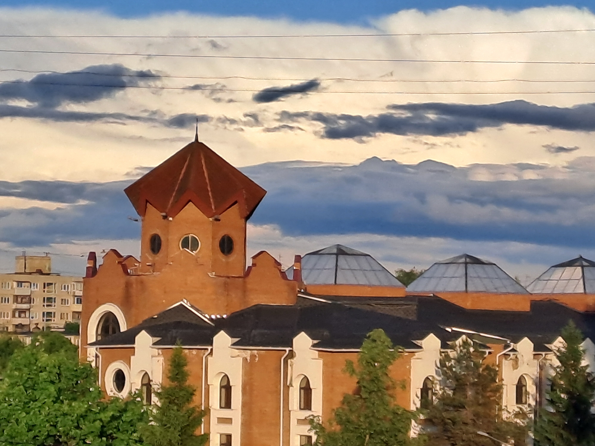 The image shows a multi-story building in Elektrostal, Russia. The building features a prominent tower with a red, multi-sided roof and circular windows. A section of the building also has dome-shaped glass skylights on a flat roof. The building's facade is a combination of brick and white architectural elements. Trees partially obscure the lower portion of the building. In the background, another multi-story residential building is visible. The sky is filled with clouds. Electrical wires are visible in the upper part of the image.
