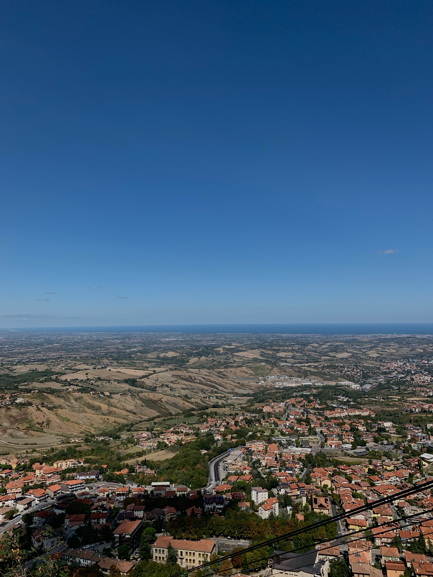 The image depicts a panoramic view of the City of San Marino, San Marino, with the Adriatic Sea visible in the distance under a clear blue sky. The foreground is dominated by a densely packed town with terracotta-roofed buildings nestled into rolling, arid hills. A winding road, bordered by cables, is visible in the lower right quadrant, suggesting a view from a cable car or a high vantage point. The mid-ground shows a spread of more urban development and agricultural land, with fields exhibiting varying shades of brown and green, indicative of dry cultivation and vegetation. The city's architecture, characterized by its red-tiled roofs and clustered buildings, stands out against the natural landscape. A faint, wispy cloud is present in the upper right portion of the sky. The overall impression is one of a historic city situated within a Mediterranean coastal environment.