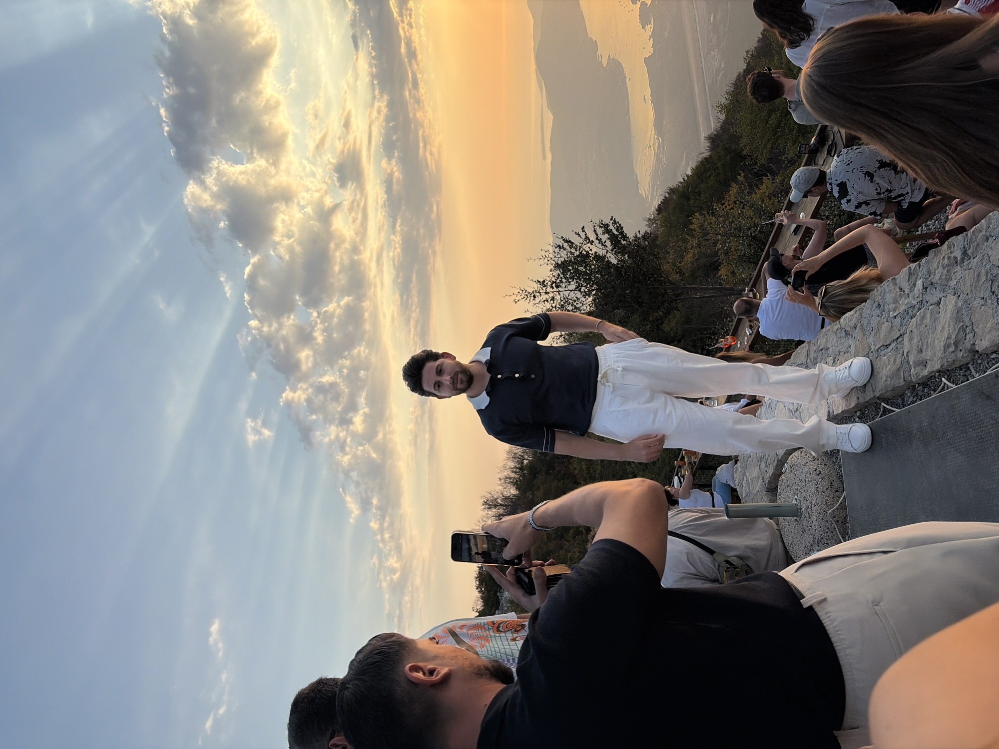 The image captures a sunset view from a high vantage point, possibly a restaurant or observation deck, in or near Kuk, Montenegro. The sky is filled with dramatic clouds illuminated by the setting sun, casting warm golden and orange hues. Beams of sunlight pierce through the clouds, creating a striking visual effect.
In the foreground, a man stands facing the camera, posing for a photo. He is wearing a dark blue short-sleeved polo shirt with a white collar and trim, and loose-fitting white trousers. He has dark hair and a beard. To his left, another person is partially visible, taking a photo of the man with a smartphone. This individual is wearing a black polo shirt and light-colored trousers.
Further in the background, several other people are seated on a tiered seating area, also overlooking the landscape. Some are holding drinks, and one person appears to be taking a photograph. The environment suggests an outdoor gathering or event, with people enjoying the scenic view.
The landscape below consists of rolling hills covered in trees, leading down to a body of water that could be a bay or a lake, with mountainous terrain visible in the distance. The lighting indicates it is late afternoon or early evening, during the golden hour. The overall atmosphere is one of leisure and appreciation of the natural beauty.