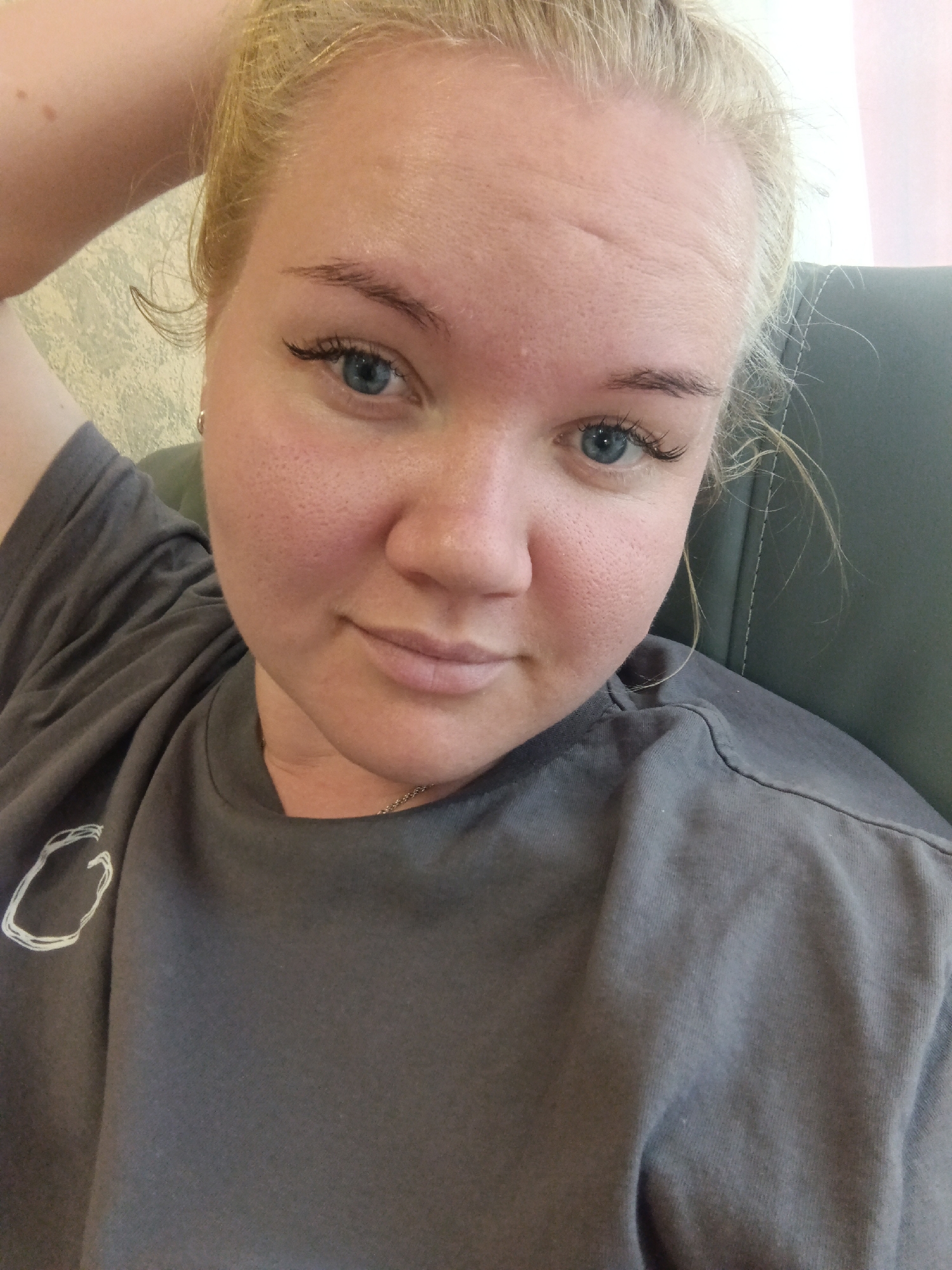 A fair-skinned, blonde woman is taking a selfie indoors, possibly in her home. She has blue eyes, fair skin with visible pores and some redness on her cheeks, and her hair is pulled up. She is wearing a dark gray t-shirt with a white circular graphic on the left shoulder. She is leaning back in what appears to be a chair, with her left arm bent and her hand behind her head. The background is blurry, showing a patterned wallpaper and the dark gray upholstery of the chair. The lighting suggests an indoor environment with natural light coming from the right. There is no indication of the location being in the city of Фоки, Russia.