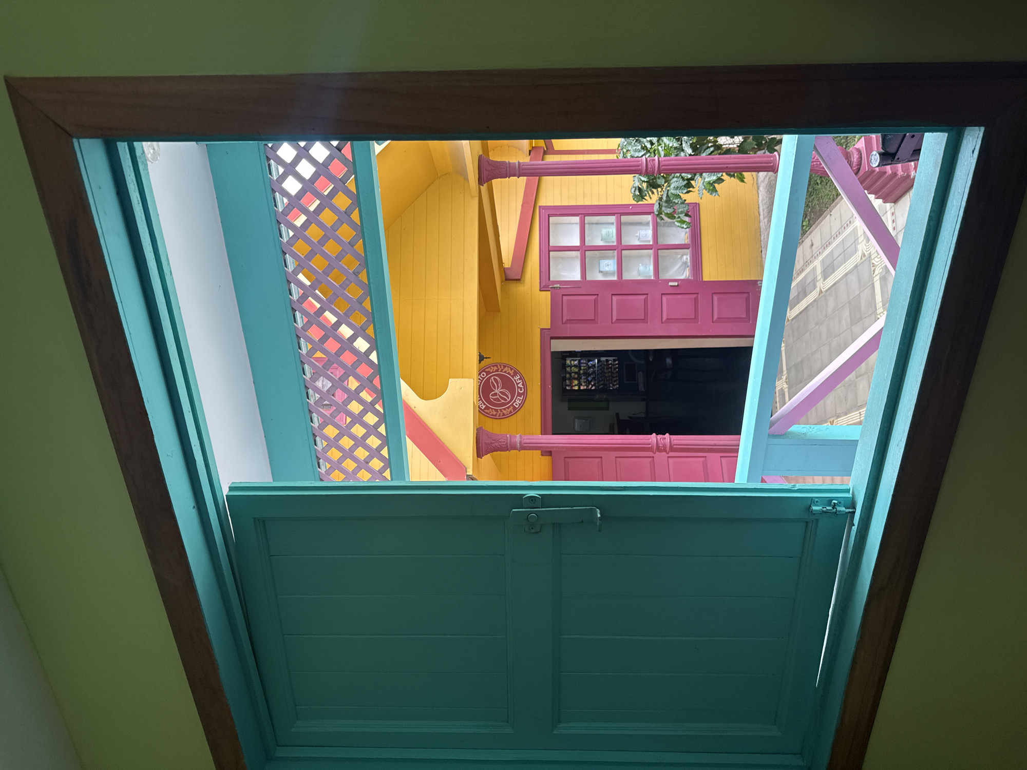 The image shows a view through an open window, looking out at a colorful building. The window frame is made of wood, with teal-colored shutters and trim. Outside, a yellow building with pink doors and window frames is visible. A sign that says "DEL CAFE" is on the building. The building has a colorful lattice roof in shades of pink, purple, and yellow. The scene appears to be in a tropical environment, with greenery visible. The image was taken in San Jose, Costa Rica, at a cafe.