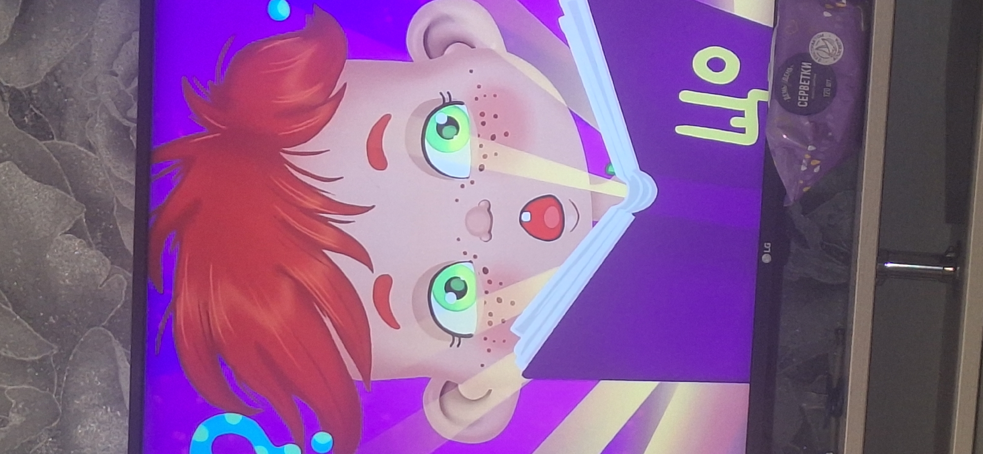 A television screen displays an animated character, a child with red hair and green eyes, with an open mouth and a surprised expression. The child is holding an open purple book, with rays of yellow light emanating from it and the child's eyes. The background is a vibrant purple. In the lower right corner of the screen, the Cyrillic letters "ЩО" are visible on the book. Below the screen, on a shelf, are a package of wet wipes labeled "СЕРВЕТКИ" and the LG logo. The television is placed on a dark, reflective surface that appears to be a piece of furniture. The wall behind the television is textured, resembling a grey stone or concrete pattern. The scene is likely indoors, in a domestic setting, as suggested by the television and furniture. There is no information in the image that places it in Bila Tserkva, Ukraine, or any other specific city.