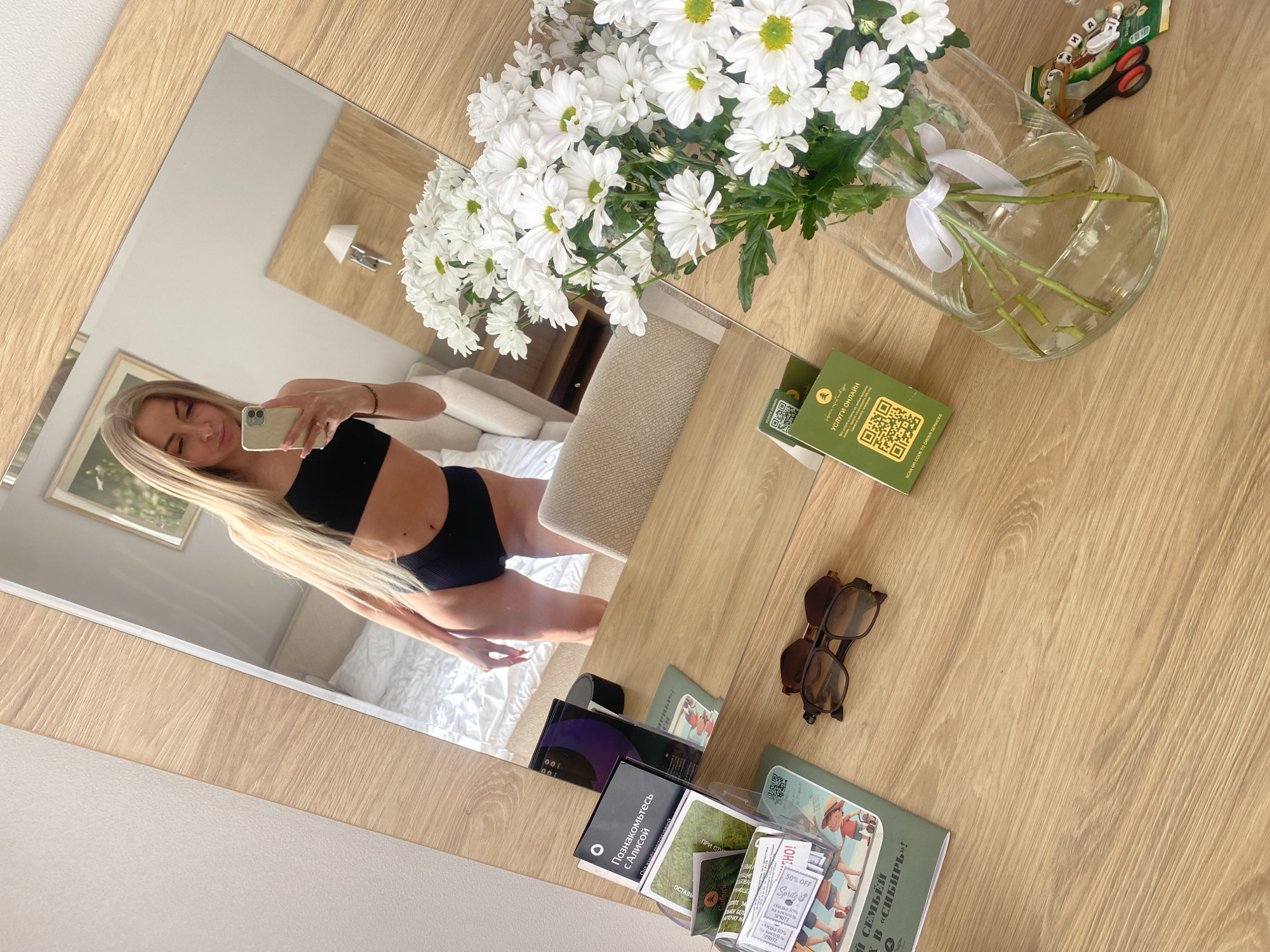 Here is a matter-of-fact description of the image:
The image shows a woman reflected in a mirror. She is wearing a black crop top and underwear and holding a mobile phone. The reflection shows a bed in a room. On the table in front of the mirror is a vase of white daisies, along with sunglasses, books in Russian, a green card labeled "УСЛУГИ ОНЛАЙН" (Online Services), scissors, and other small objects. The text on the books suggests the location could be Tyumen, Russia.