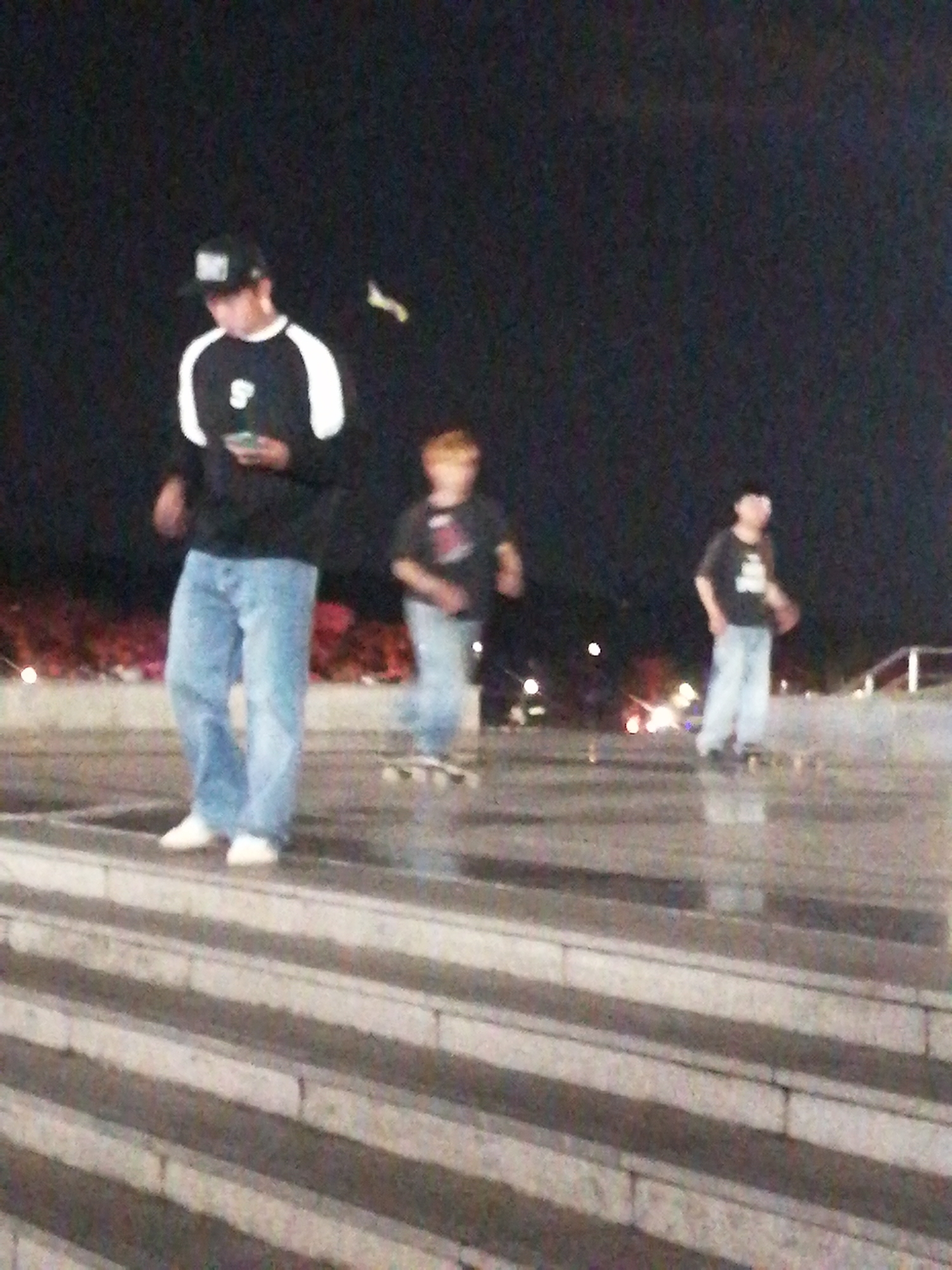 In the nighttime, three young males are in focus, engaged in skateboarding on a set of broad, concrete steps. The foremost individual, wearing a black baseball cap and a black and white raglan shirt with blue jeans, is holding a phone in his left hand. Behind him, another male in a black t-shirt is partially visible. Further back, a third male, also in a black t-shirt and light-colored pants, is seen skateboarding. The background is dark, with a faint glow of lights from what appears to be a distant urban area, suggesting this scene is situated within the city of Da Lat, Vietnam. The lighting is artificial, likely from streetlights or the ambient city glow, creating a moody atmosphere.
