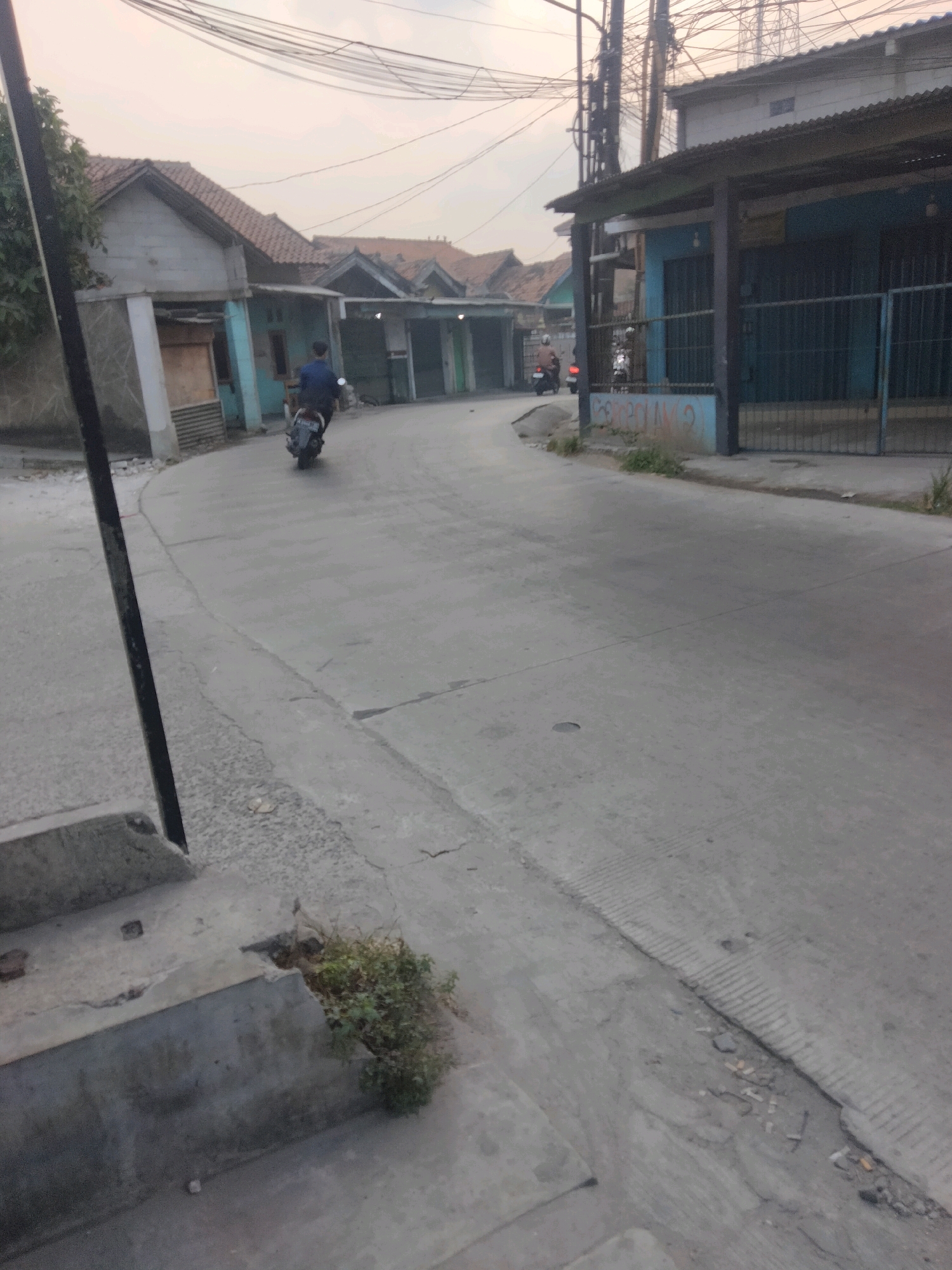 The image depicts a street scene in Pisangan Jaya, Indonesia. A person is riding a motorcycle along a paved road that curves to the right. The road is flanked by buildings and vegetation. On the left side of the road, there are houses with tiled roofs and some are painted. A dark pole is visible on the far left foreground. On the right side of the road, there are more buildings, some appearing to be shops with covered entrances and metal gates. The sky above is overcast. There are power lines crisscrossing the upper portion of the image. The overall impression is that of a residential and commercial street in an urban or semi-urban area.