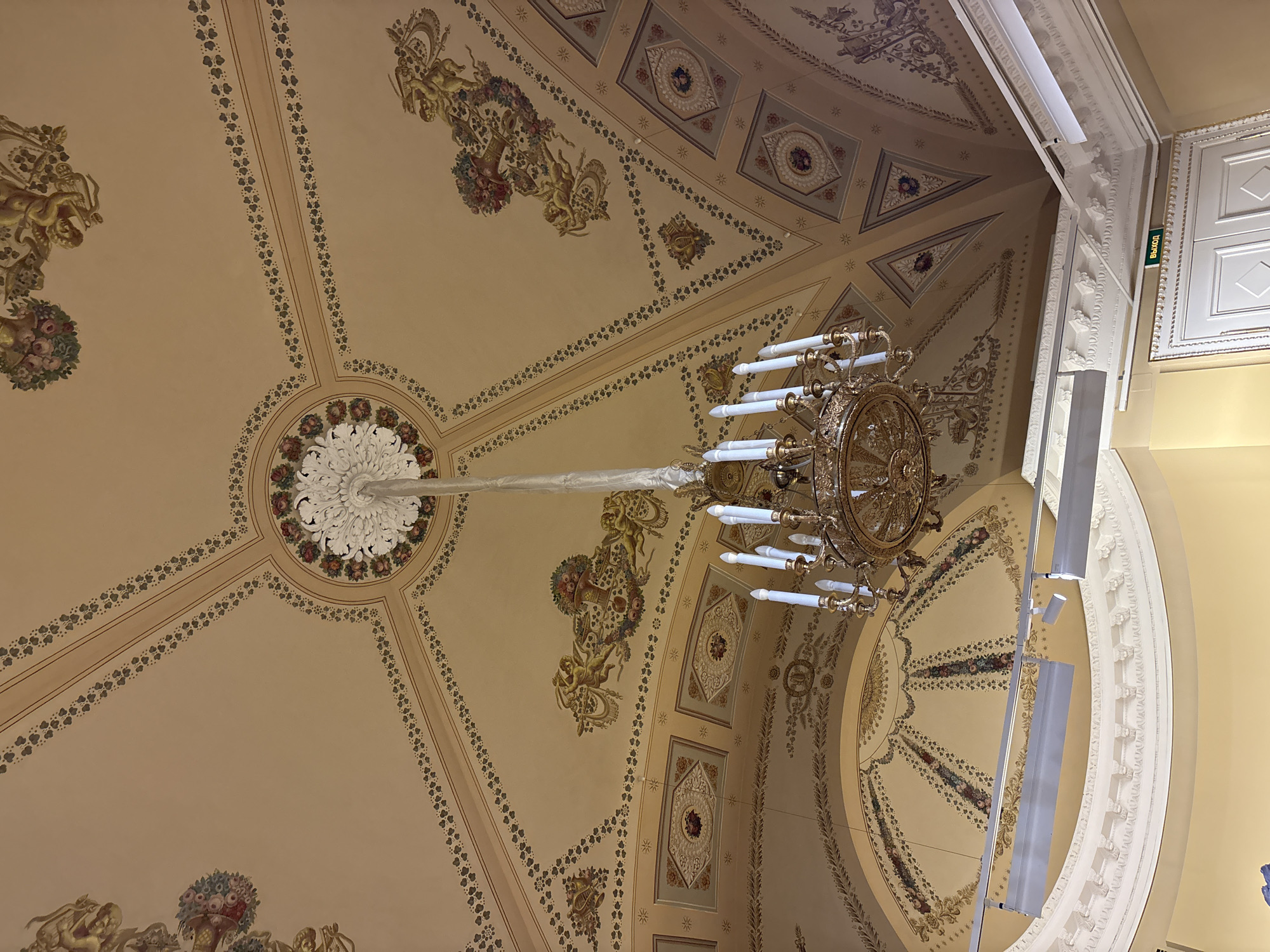 This image shows a detailed view of a ceiling inside a building in Saint Petersburg, Russia. The ceiling is adorned with intricate paintings and decorative elements, including patterns, floral designs, and depictions of cherubic figures. A large, ornate chandelier hangs from the center of the ceiling, adding to the opulence of the space. The architecture reflects a classical or neoclassical style, characterized by symmetry and refined aesthetics. The room appears to be well-lit, showcasing the ornate details of the ceiling. The presence of an "exit" sign and what appears to be a modern lighting fixture indicate that the building is in use.