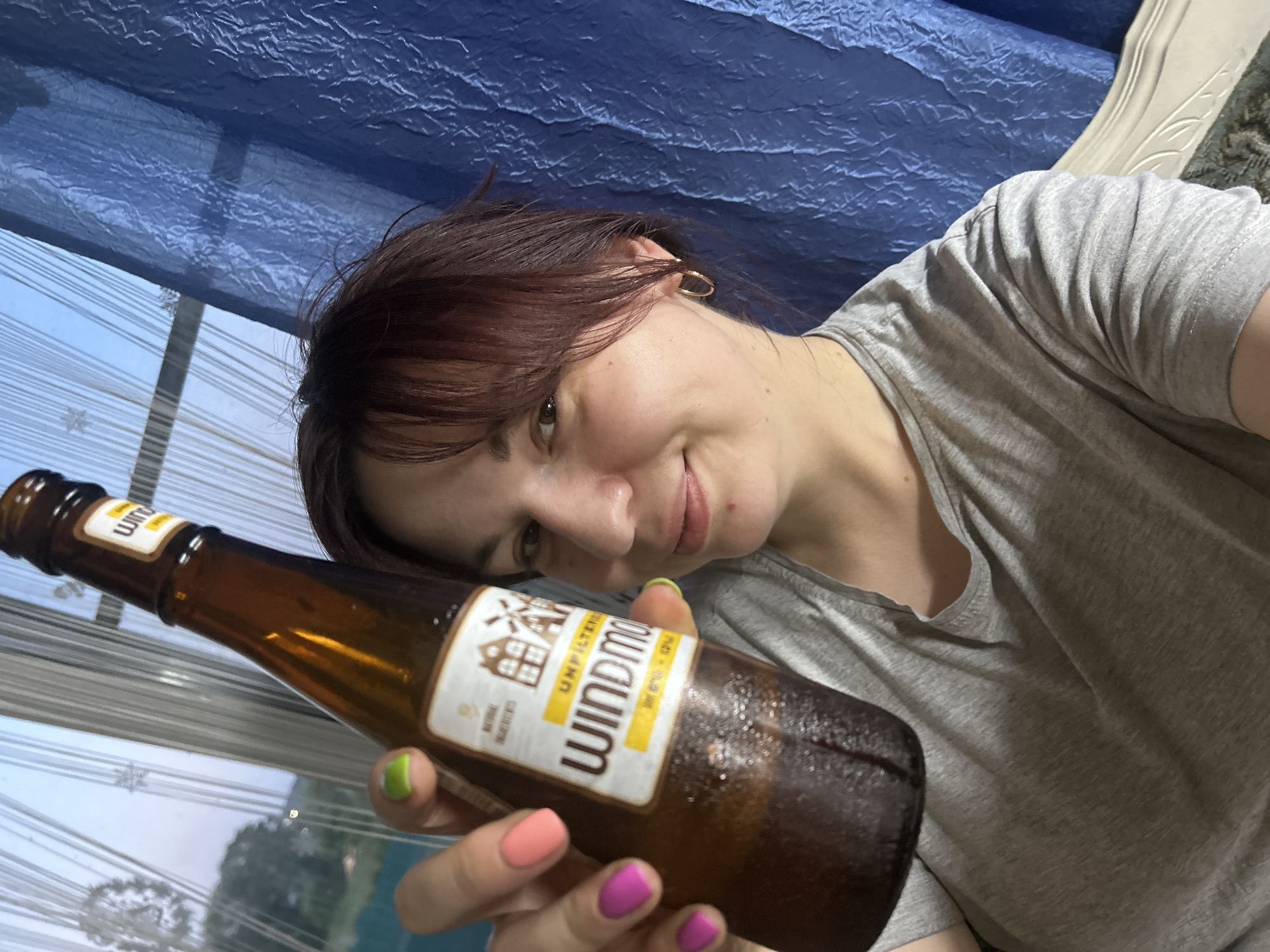 A woman is holding a bottle of Windmill unfiltered beer up to the camera. She is wearing a gray t-shirt and has brightly colored nail polish on her fingers. The background shows a window with blinds, a blue curtain, and some greenery outside. The scene appears to be indoors, possibly in a home.