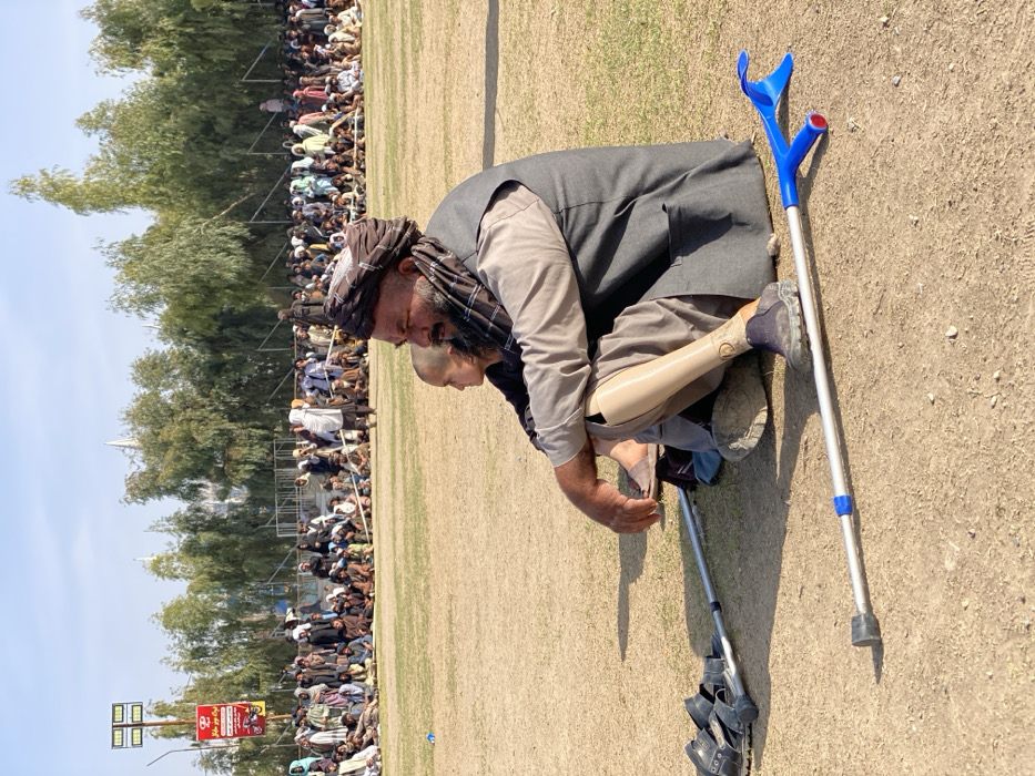 The image captures an outdoor scene in Kandahar, Afghanistan, at what appears to be a public field or stadium. In the foreground, an adult male with a dark beard and turban is seated on dry, light brown ground with sparse grass, facing towards the right. He wears a light-colored tunic, a dark sleeveless vest, and has a visible beige prosthetic left leg, adorned with a dark shoe. His right foot, in an open-toed sandal, is bent. A young child, with a shaved head and dark clothing, sits immediately behind and to the left of the man, also looking in the same direction. Two crutches are laid horizontally on the ground in front of them: one silver underarm crutch with black rubber fittings, and one silver forearm crutch with a blue cuff and handgrip. A black sandal rests on the underarm crutch.
In the middle ground, a large gathering of individuals, primarily adult males in traditional attire, are seated or standing along a perimeter line marked by a white rope or barrier on a more densely grassy area. They form a wide arc across the frame, observing an event outside the camera's view. A line of tall, slender trees with light green foliage forms a backdrop behind the crowd.
The background shows a clear blue sky. To the far left, a tall light pole equipped with multiple floodlights stands beside a red and white billboard featuring text in Dari/Pashto, including "کندهار" (Kandahar) and "نوروز مبارک" (Happy Nowruz), along with images of a motorbike and a car. Further in the distance, partially obscured by the trees, the white spires of a large building are visible.