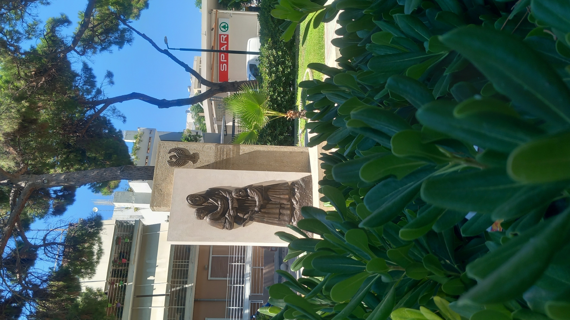 This is a low-angle, vertical, full shot of a memorial sculpture on the island of Kos, Greece. The sculpture features a bronze relief of a woman holding a child, with another child standing beside her. Behind the relief, the memorial is constructed of stone. To the right of the sculpture, a small palm tree is visible. In the background, a white building with balconies and potted plants can be seen. Further in the background, a Spar supermarket with a white van and a streetlight are present. The foreground is dominated by lush green foliage, obscuring the lower portion of the memorial and the ground. The sky is a clear blue. The memorial is located in the city of Kos, Greece, specifically in the vicinity of the Spar supermarket.