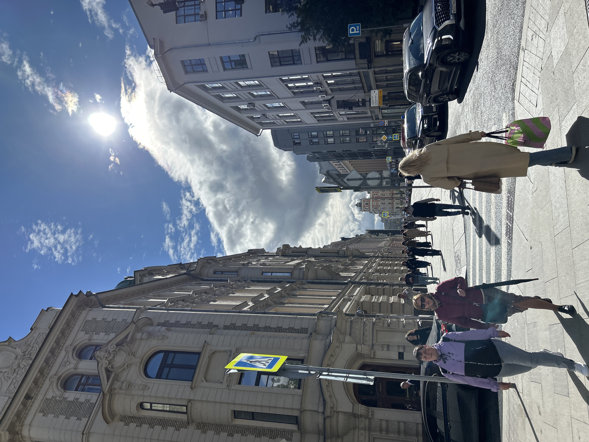 The image shows a street scene with people walking on the sidewalk. Buildings line the street on both sides, with cars parked along the curb. A pedestrian crossing sign is visible. The sky is blue with clouds and the sun shining brightly. The location is a city.