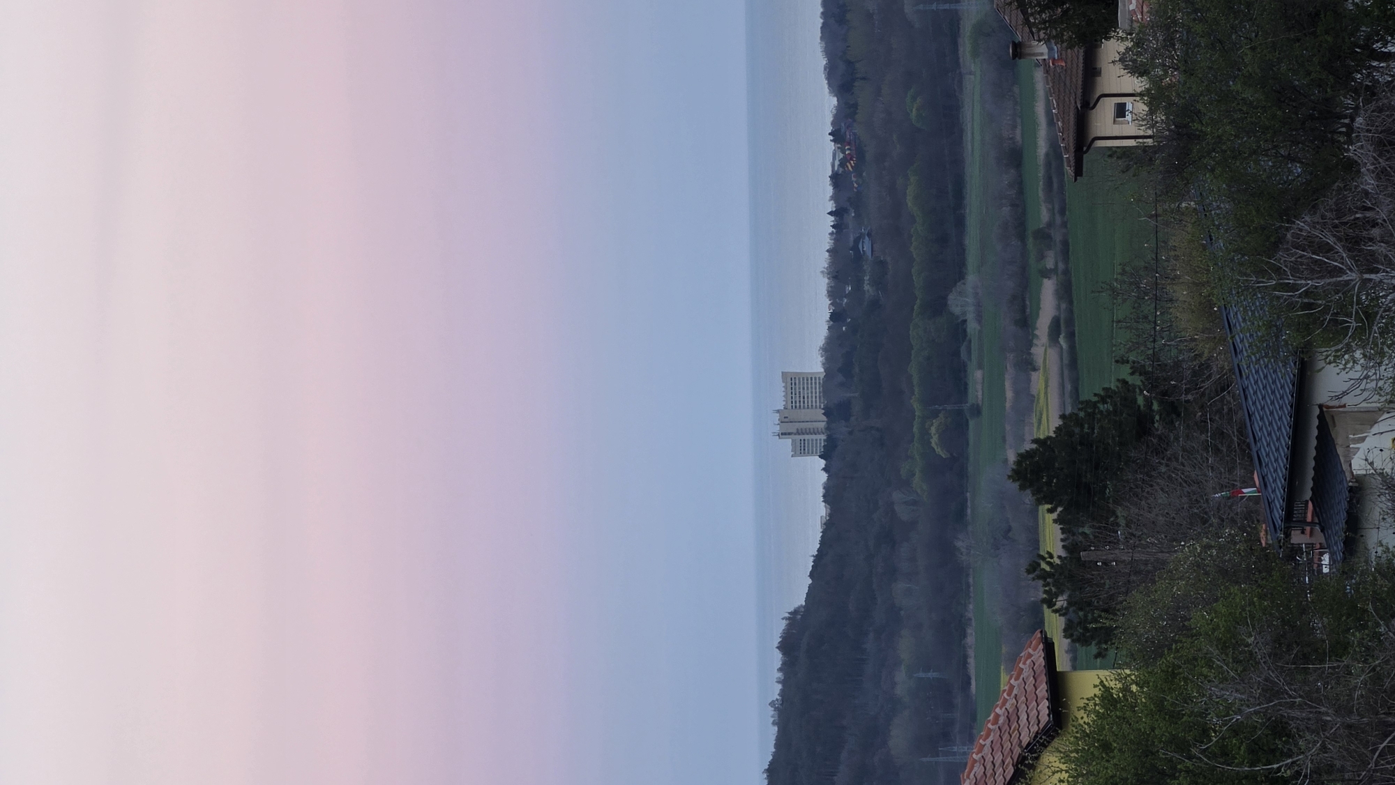 A landscape view from Rogachevo, Bulgaria, featuring a building on the coastline, a green valley, and residential areas. The sky transitions from a soft pink to light blue. The building on the coastline is a high rise apartment building. Trees in the valley are bare of leaves.