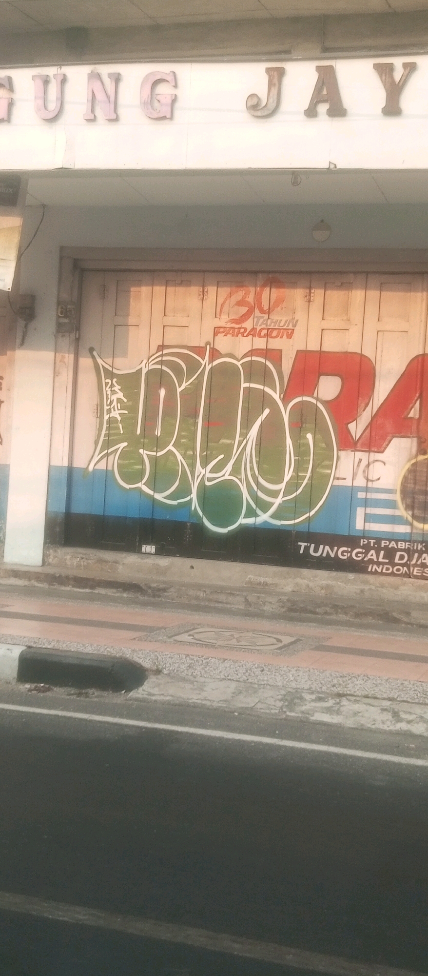 The image captures a street scene in Kediri City, Indonesia, featuring a building with a large garage door that has been painted with graffiti. Above the garage door, the words "GUNG JAY" are visible in large, faded letters, indicating the name of the establishment. The garage door itself is wooden and has graffiti covering a significant portion of it. The graffiti includes large, stylized lettering in green with white outlines, and a red stylized numeral "30" with the text "TAHUN PARAGON" underneath. Smaller text, possibly a company name, is also present on the lower right side of the garage door, reading "PT. PABRIK TUNGGAL DJA INDONESIA". The surrounding environment shows a sidewalk with decorative paving stones and a section of asphalt road in the foreground. The lighting suggests it is daytime, possibly late afternoon due to the long shadows. There are no people visible in the image.