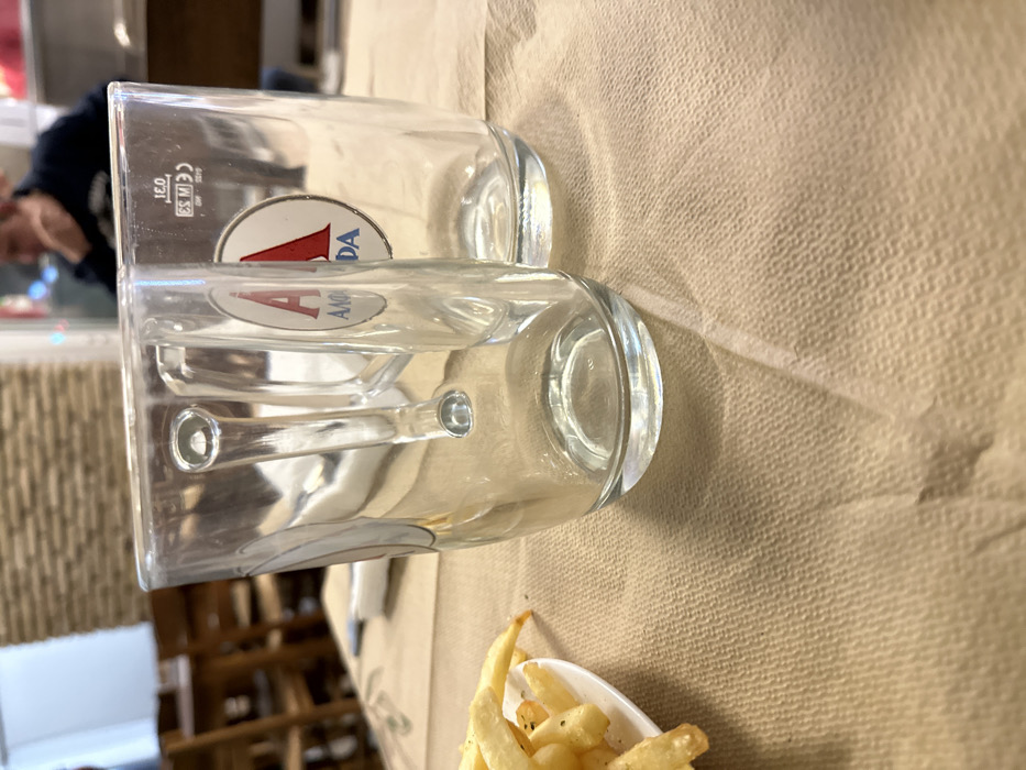 The image displays two clear glass cups resting on a light brown, textured surface, likely a disposable paper tablecloth, in what appears to be an indoor setting like a restaurant or bar. The image is oriented sideways, with the cups lying horizontally.
In the foreground, towards the bottom right, a small white bowl contains a serving of golden-brown french fries, seasoned with visible herbs (possibly parsley or oregano) and black pepper.
The two clear glass cups are positioned centrally. Both are empty. The top cup, which is slightly behind the bottom cup, features a white circular logo with a prominent red letter "A" and the word "AMSTEL" or "AMANDA" (appears to be "AMSTEL" based on typical branding, with "AMANDA" also visible) written in blue underneath it. Etched markings are visible on the side of this cup, including "0.3 L" (indicating a 0.3-liter capacity), "CE M 59", and "4426". The bottom cup, slightly in front, also bears the same "AMANDA" logo. This cup has two distinctive raised, circular features on its side, possibly for grip or as a design element.
The background is softly blurred, providing context to the scene. On the far left, a person's head and shoulders, wearing a dark top, are faintly visible, out of focus. The background also shows hints of an interior space with wooden structures, possibly shelves, tables, or a bar counter, and a textured wall with vertical patterns. The lighting suggests an indoor environment, creating soft shadows cast by the cups and the fry bowl on the tablecloth.
The overall scene depicts a casual dining or drinking atmosphere, possibly during a meal or a break, with beverages (indicated by the glasses) and a snack of french fries.