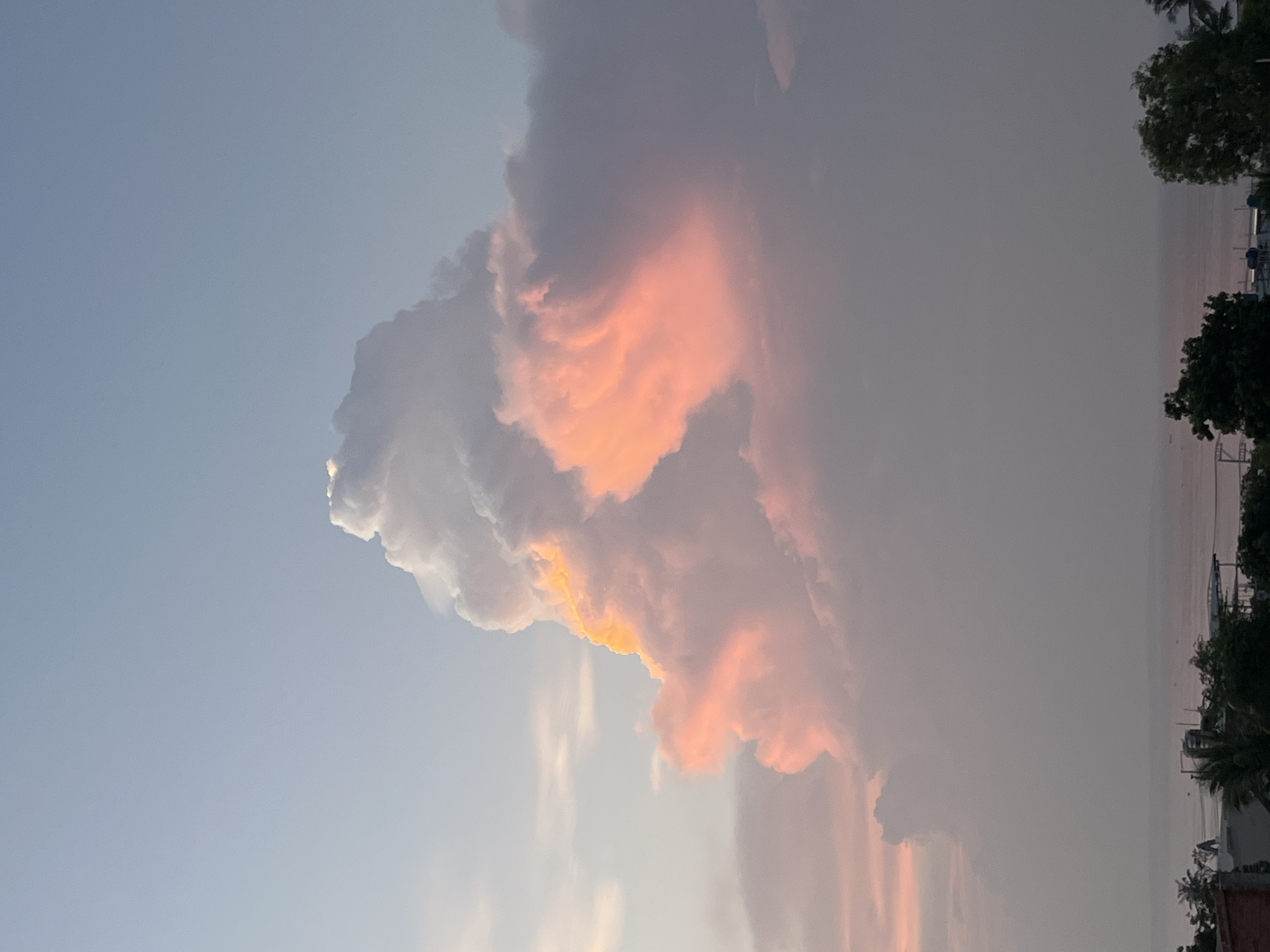 A dramatic sunset casts a warm, pink and orange glow on a towering cumulonimbus cloud formation, creating a striking contrast against the pale blue sky. The cloud, occupying the center and right portions of the frame, exhibits intricate textures and shadows, with its upper peak catching the brightest light. Below the sky, a muted horizon reveals the ocean, its surface reflecting the soft twilight hues. Along the shoreline, silhouetted against the dim light, are the outlines of trees and buildings, suggesting the coastal city of Mũi Né, Vietnam. There are no visible people or animals in the image. The overall impression is one of a serene yet powerful natural spectacle.