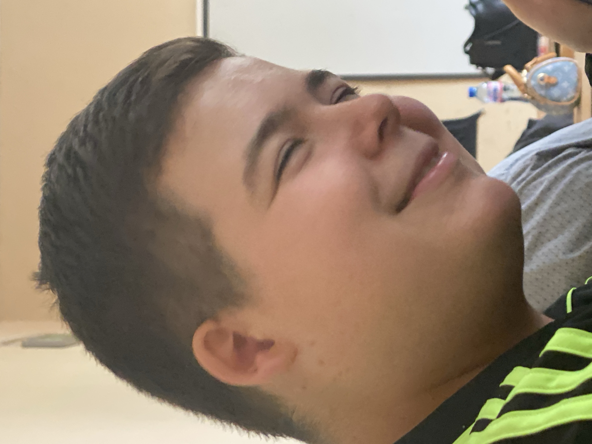 A close-up shot depicts a person's face in profile, lying down. The individual appears to have short, dark hair and is fair-skinned. Their eyes are closed, and a slight smile is visible. They are wearing a black shirt with bright green stripes. The background is blurred and indistinct but shows a white board on the wall, and other objects including a water bottle. The location is Unknown, Unknown.
