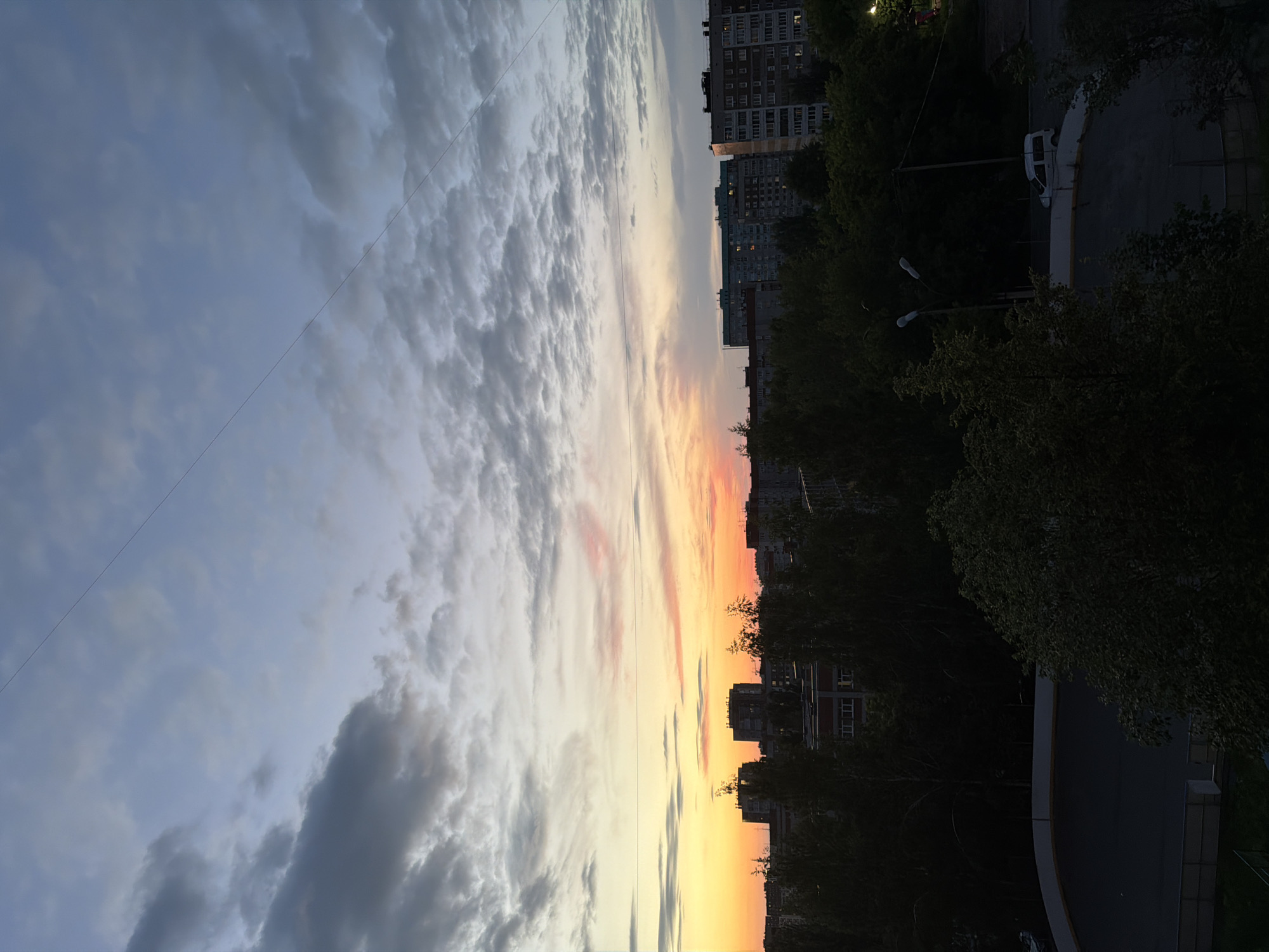 The image depicts an urban landscape at dusk, likely in Izhevsk, Russia. The sky is a blend of pale blue and grey clouds, with a band of warm orange and pink hues near the horizon, indicating sunset. Silhouetted against the sky are several multi-story apartment buildings of varying architectural styles, typical of Soviet-era urban planning. Dense green trees form a dark layer in the foreground, obscuring the lower portions of the buildings and the ground. A thin, dark line, possibly a power line or wire, bisects the upper portion of the sky diagonally from left to right. In the lower part of the image, a dark paved area, possibly a sports court or playground, is visible, bordered by a low fence. A white van is parked on the paved area. The scene is quiet and lacks any visible human activity.