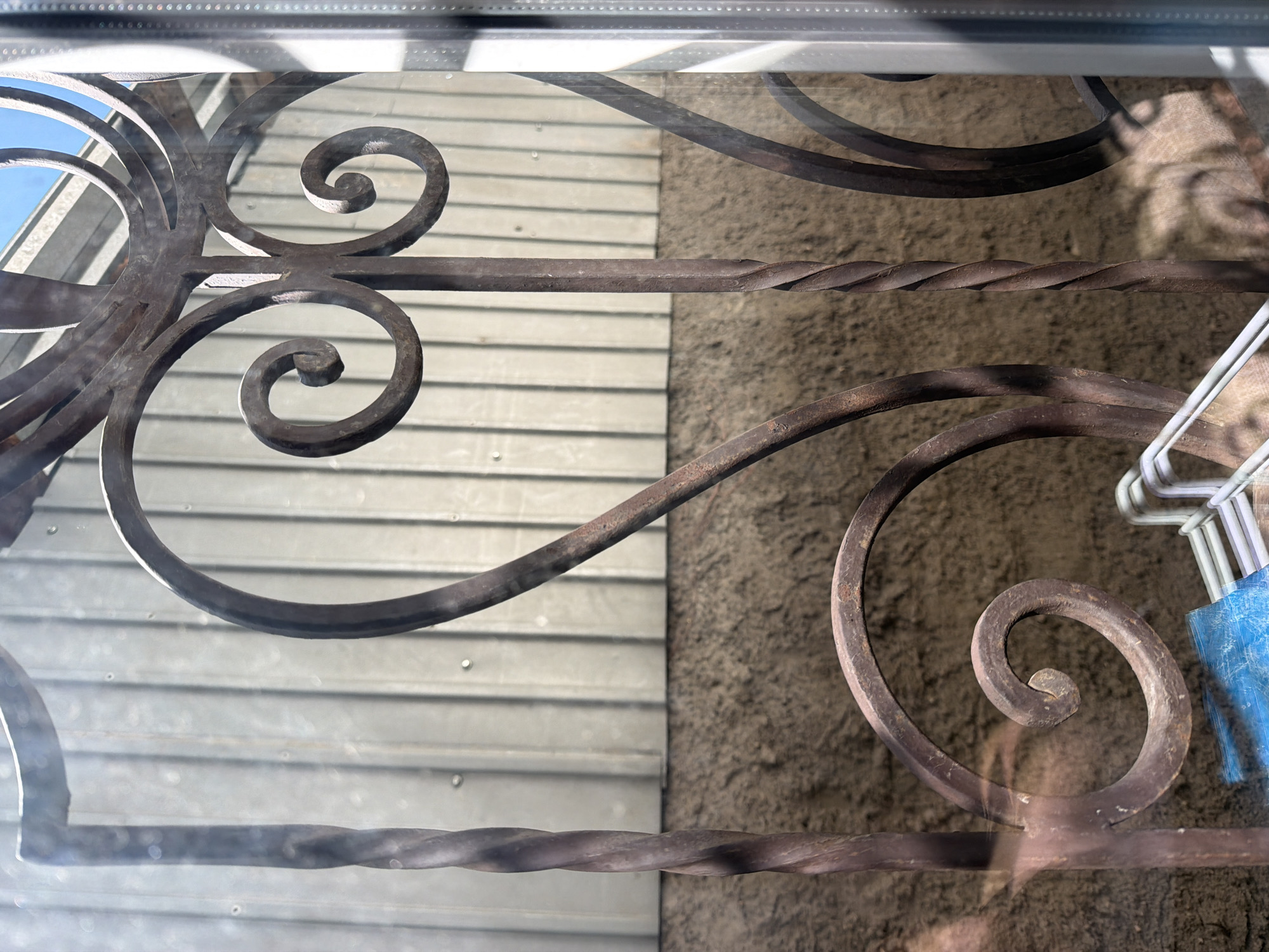 The image displays a close-up view of a rustic, wrought-iron gate or fence with decorative scrollwork. The metal has a reddish-brown, weathered patina, suggesting age and exposure to the elements. The gate features twisted metal elements and curved designs, including prominent spiral shapes. Behind the gate, a corrugated metal wall is visible, with vertical ribs. Below this wall, there is a textured surface, possibly stucco or rough-cut stone. Sunlight casts shadows and highlights on the metalwork, creating a play of light and dark. In the lower part of the frame, parts of white clothes hangers and a blue object are visible, partially obscured by the gate. The image is taken through a reflective surface, likely a window, as evidenced by faint reflections in the upper left corner and a vertical glare on the right edge. The overall impression is one of an outdoor scene, potentially a garden, yard, or an entrance. The location is specified as the city administration of Qarağandı city, Kazakhstan.