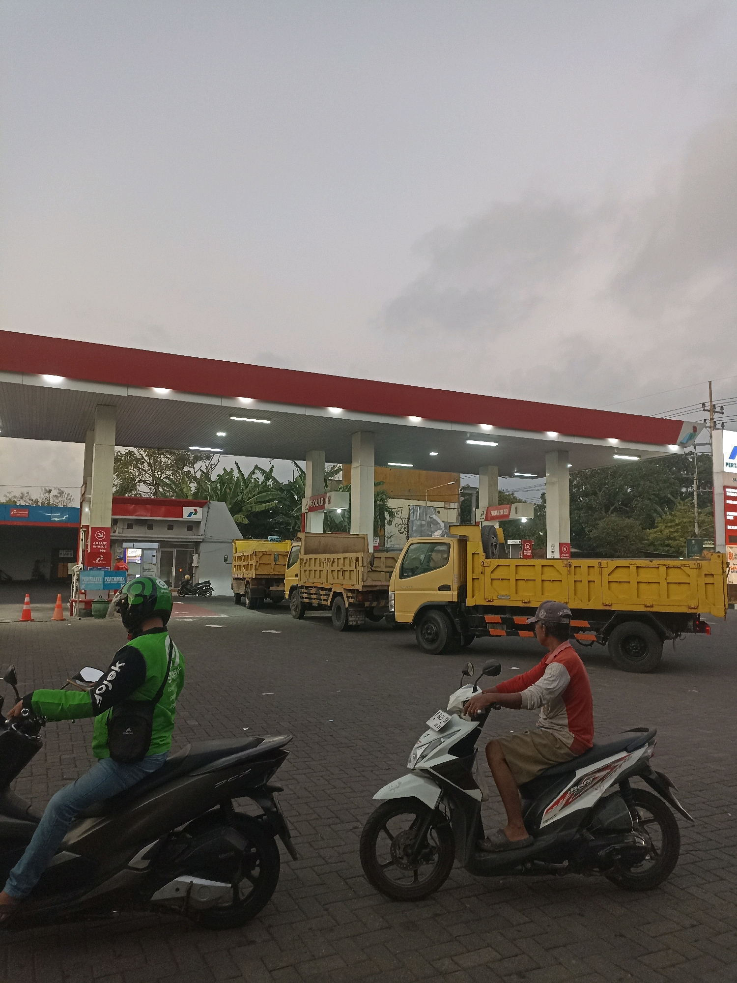 This is a forensic, matter-of-fact description of the image:
The image depicts a scene at a Pertamina gas station in Mojopanggung, Indonesia, likely during overcast or dusk conditions. In the foreground, two individuals are riding motorcycles. The person on the left is wearing a bright green jacket with the "Gojek" logo and a green helmet. They are on a dark-colored scooter. The person on the right is wearing a red and white long-sleeved shirt and a cap, riding a white scooter.
Behind the motorcycles, under the covered area of the gas station, are several large yellow dump trucks. The gas station has a red roof supported by white pillars, and various signs and pumps are visible. Trees and foliage are present in the background, suggesting an outdoor setting. The ground is paved with interlocking blocks. In the distance, a building with a Pertamina logo is visible. Orange traffic cones are placed on the ground near the gas pumps. The sky is predominantly gray and cloudy.