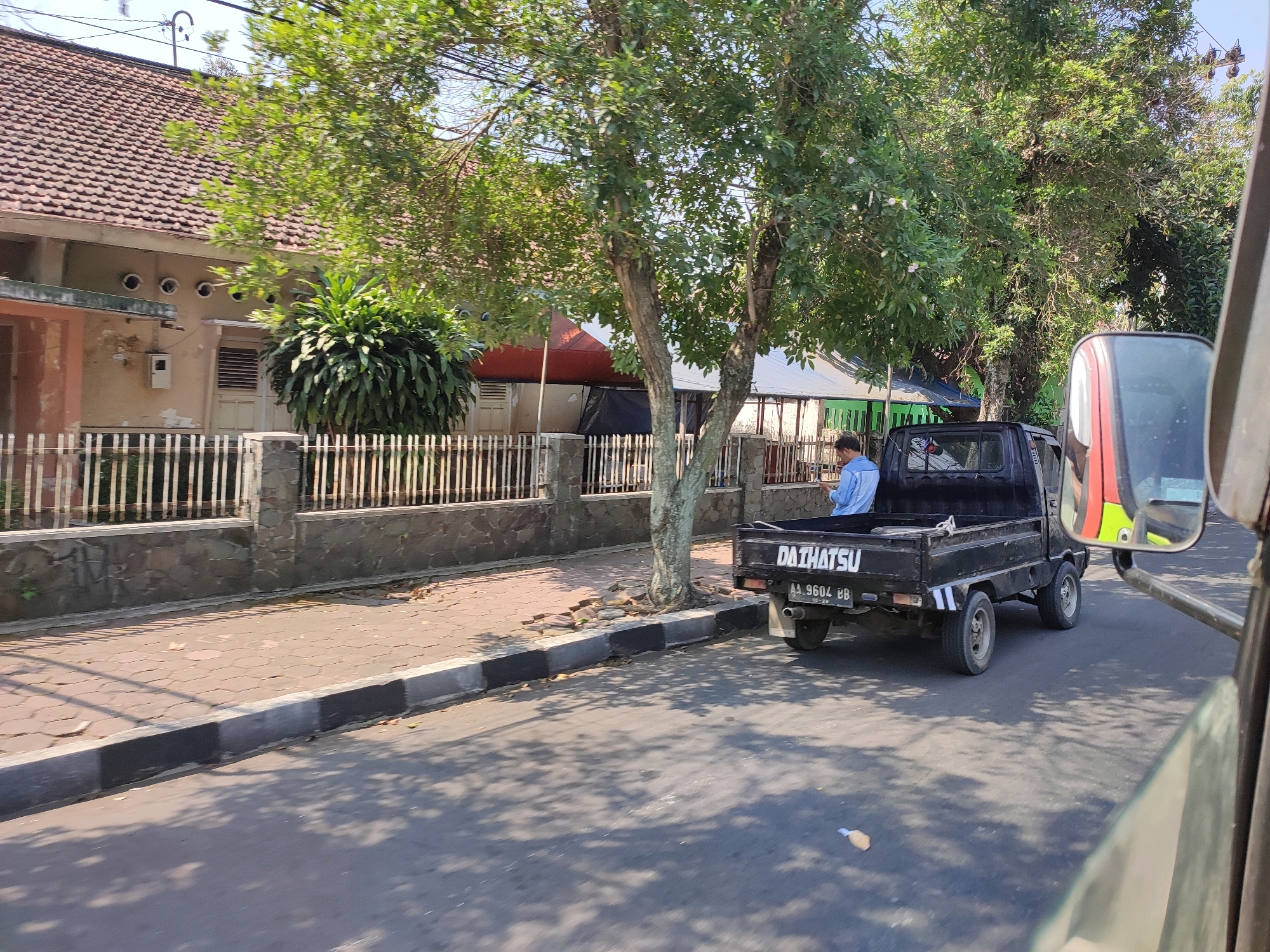 The image captures a street scene in Magelang, Indonesia, on a sunny day. A black Daihatsu pickup truck is parked on the right side of the road. A person in a blue shirt is sitting in the truck bed. The truck has a license plate that reads "AA 9604 BB".
On the left side of the road, there is a sidewalk made of interlocking concrete blocks. Beyond the sidewalk is a low fence made of vertical wooden planks and a stone wall base. Behind the fence, a building with a tiled roof is visible, featuring several small circular windows and a beige-colored wall. A lush green bush grows near the building. Trees with dense foliage line the street, casting shadows on the road and sidewalk. The overall environment suggests a residential or commercial area within the city.
