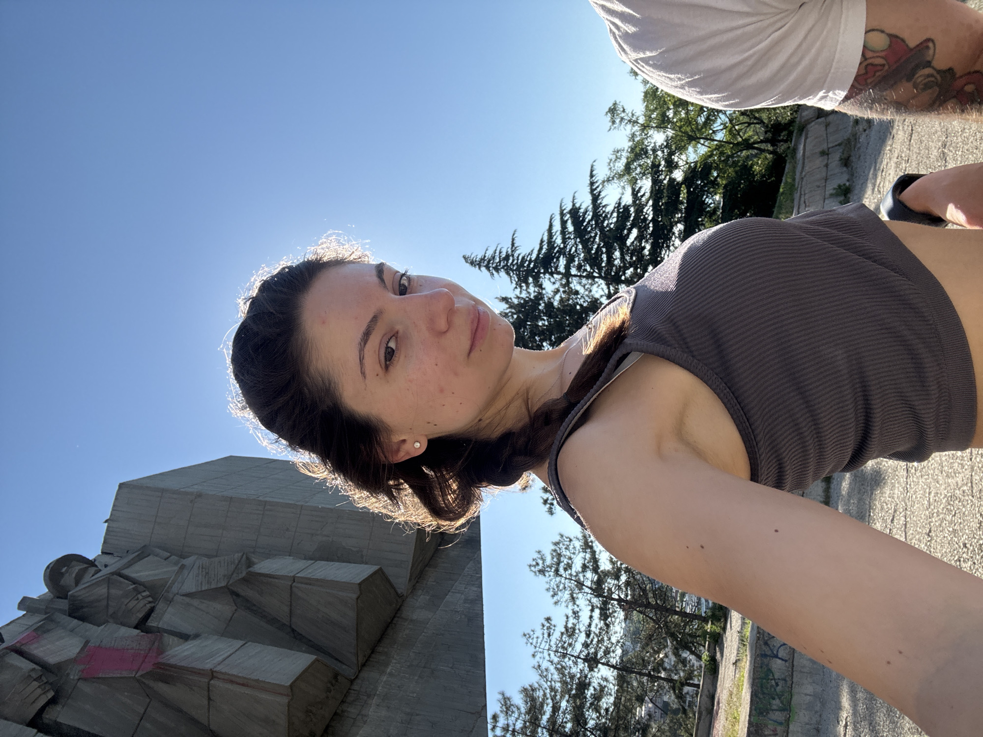 A young woman with dark hair braided and a grey tank top poses for a selfie in front of the Monument of the Bulgarian-Soviet Friendship in Varna, Bulgaria. The monument is a large, abstract concrete structure with geometric shapes, some of which are marked with pink paint. The woman is positioned in the foreground, with the monument and a bright blue sky in the background. Trees are visible behind the monument, and another person's tattooed leg is partially visible on the right side of the frame. The lighting suggests it is daytime, with the sun highlighting the woman's hair and the top of the monument.