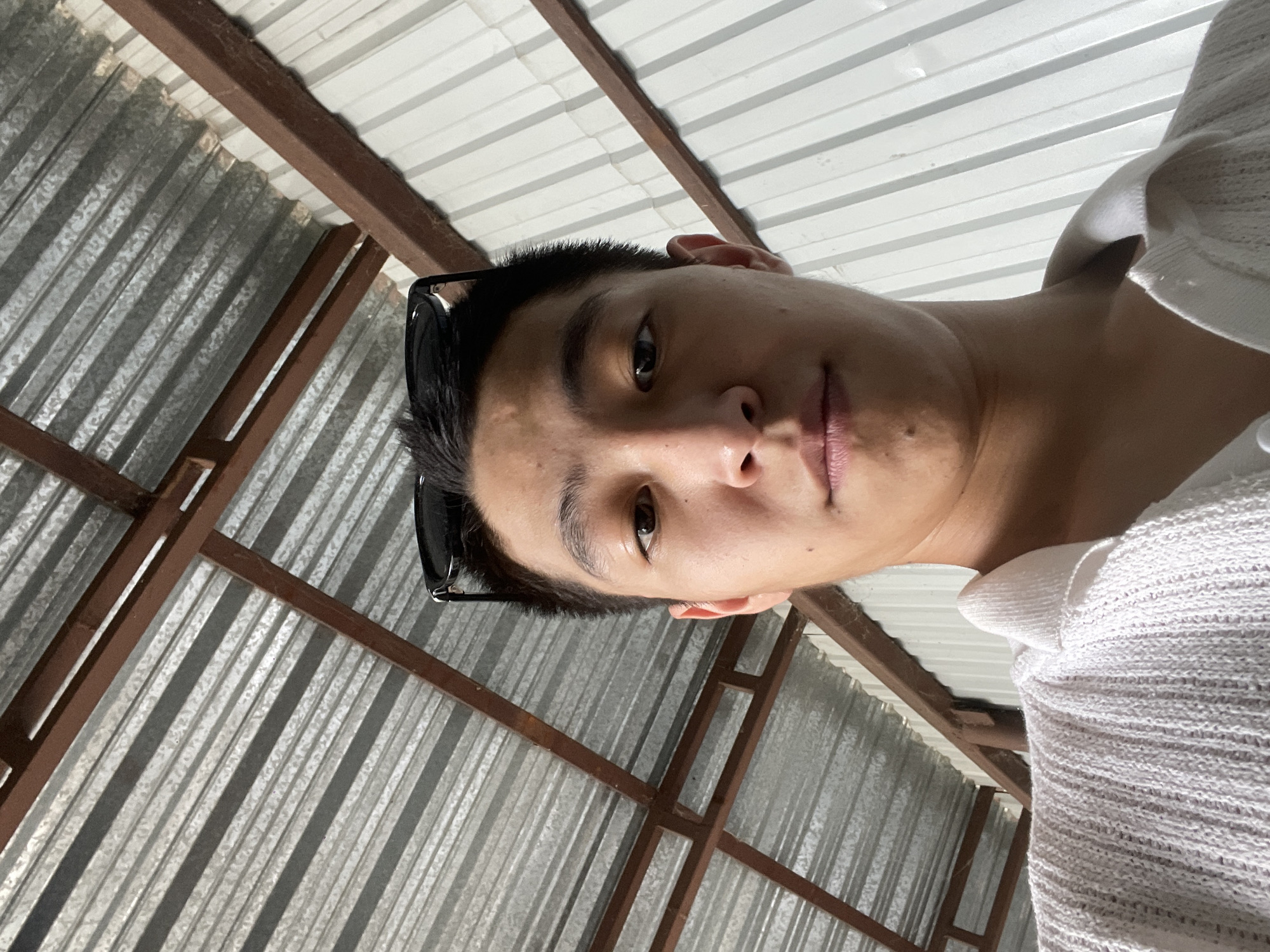 The image shows a close-up, low-angle selfie of a young man wearing a white collared shirt. He has sunglasses perched on his head. The background consists of a corrugated metal roof with visible brown metal supports, and a white corrugated metal wall. The scene appears to be outdoors, possibly under a covered structure. The lighting suggests it is daytime. The location is specified as Кенен Әзірбаев ауылы, Kazakhstan.
