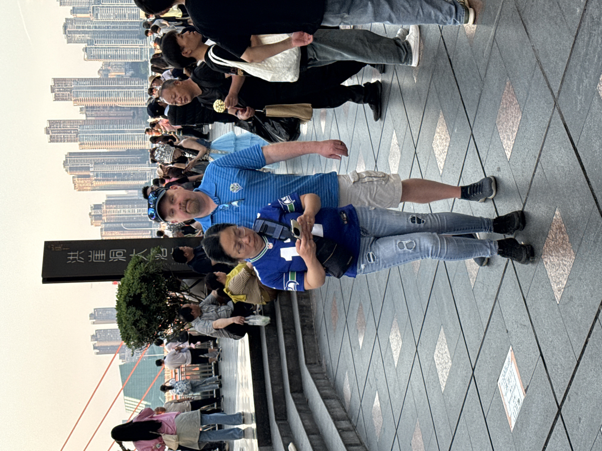 A medium shot shows a crowd of people on an overcast day in the Yuzhong District of China. The scene is along a paved area with a diamond pattern, with buildings in the background. A sign is visible on the left side. In the foreground, a man with sunglasses on his head wears a blue polo shirt and khaki shorts, standing next to a woman wearing a blue Seahawks jersey and ripped jeans, holding a phone. The crowd is diverse, including people of different ages and ethnicities. The subjects are generally standing, looking at their phones, or looking at the scenery.