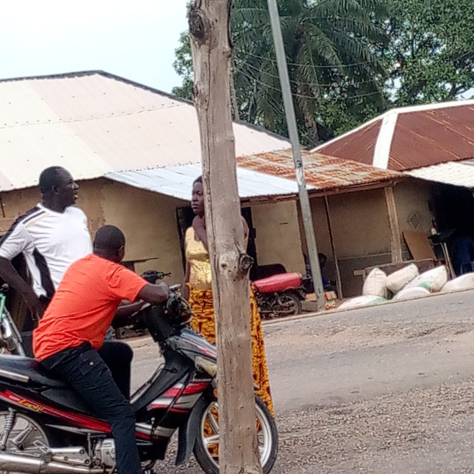 The image depicts an outdoor scene on a road in Mararaba, Nigeria. Three people are visible: two men and a woman. One man is wearing a white shirt with black sleeves and is looking and talking to the man who is on a black and red motorcycle and wearing an orange t-shirt and black pants. A woman wearing a gold top and patterned yellow and orange skirt stands behind a thick wooden pole.
Behind them are several shacks with corrugated metal roofs. There are several bags of what appears to be grain or other agricultural product stacked near one of the shacks. Another motorcycle is parked near one of the shacks. A utility pole is visible on the right side of the image. The road appears to be made of asphalt and is in rough condition. Palm trees are visible in the background.