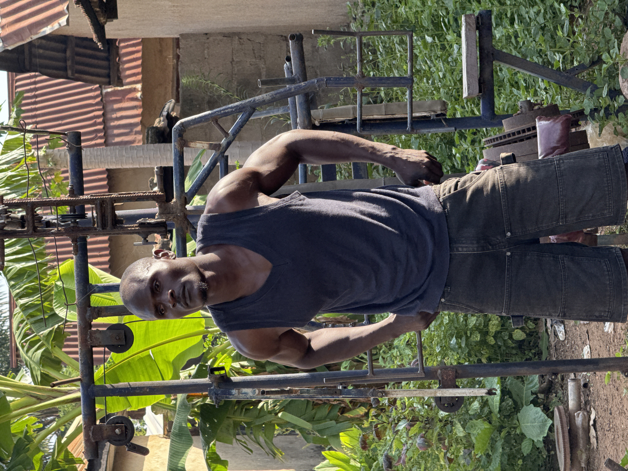 A dark-skinned man with a shaved head and a goatee stands in front of a homemade weightlifting machine in Akwanga, Nigeria. He is wearing a dark grey V-neck tank top and dark grey denim shorts. The weightlifting machine is made of metal pipes and pulleys, and appears to be rusted and old. There are weights and other metal parts around the machine. Behind the man, there are banana trees, buildings with corrugated metal roofs, and other vegetation. The ground is dirt and there is some litter visible. The scene appears to be outdoors in a somewhat rural or undeveloped area.
