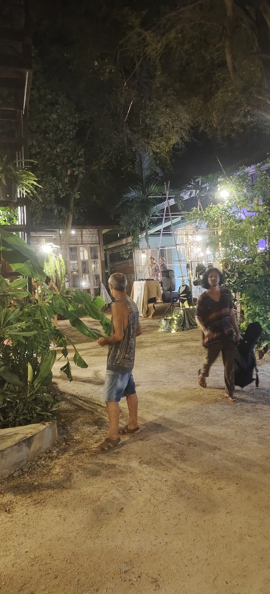A wide shot captures an outdoor nighttime scene in a dimly lit area that appears to be a courtyard or garden space in Ko Pha-ngan District, Thailand. In the foreground, a man with short grey hair, wearing a dark sleeveless shirt with a graphic print and denim shorts, stands facing away from the camera, holding large green leaves. He is wearing brown sandals. To the right, further back, another person, wearing dark clothing, is walking and carrying a black bag that could be a guitar case. They are also wearing sandals. In the midground, there is a raised platform with a table covered by a patterned cloth, two speakers, and a bamboo structure with lights. Two figures appear to be standing behind the table, possibly performing or setting up equipment. The ground is a light-colored gravel or dirt path. Lush green foliage is visible on the left and right sides of the frame, and trees with dark leaves form a canopy overhead, illuminated by scattered yellow lights. Structures with wooden elements and possibly windows are visible in the background. The overall atmosphere suggests a casual outdoor gathering or event.