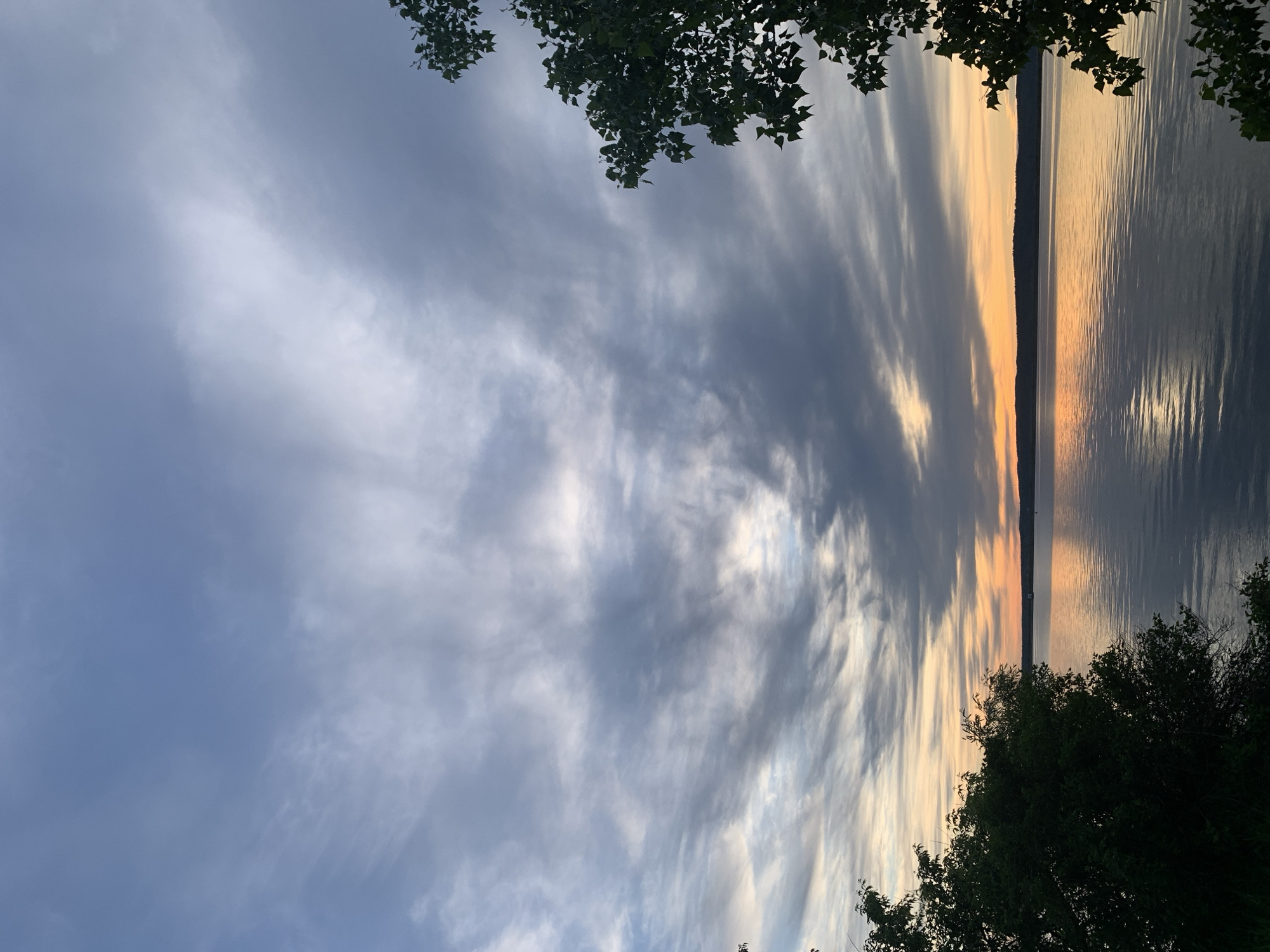 The image captures a sunset over the Kama River in Chaikovsky, Russia. Wispy clouds dominate the sky, reflecting the colors of the setting sun. The river surface is mostly calm with light reflecting from the sun. Silhouetted tree foliage frames the top and bottom of the image, providing a natural foreground.
