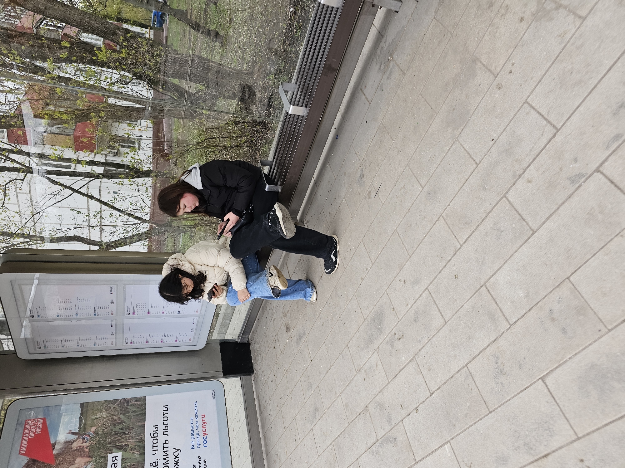 Two young women are captured in an outdoor urban setting in Moscow, Russia, possibly at a bus stop or public waiting area. Both are engrossed in their mobile phones.
The woman on the right wears a black puffer jacket with a grey hood, black pants, and white sneakers with black accents. She stands near a dark metal slatted bench. The woman on the left is dressed in a cream-colored puffer jacket, light blue jeans, and light sneakers. She leans casually against a modern public information display or bus stop shelter.
The ground is paved with light-colored rectangular tiles. In the background, trees with emerging green leaves suggest it is springtime. Through the trees, residential buildings are visible. The sky appears overcast, indicating a mild, cloudy daytime.
Visible text on an advertising panel includes "НАЦИОНАЛЬНЫЕ ПРОЕКТЫ РОССИИ" (National Projects of Russia) and phrases such as "Всё решается проще, чем кажется. госуслуги" (Everything is simpler than it seems. Gosuslugi), referring to the Russian government services portal. Another partial phrase, "чтобы омить льготы" (to apply for benefits), is also visible. The scene portrays a common urban moment of individuals occupied with their devices while waiting or resting.