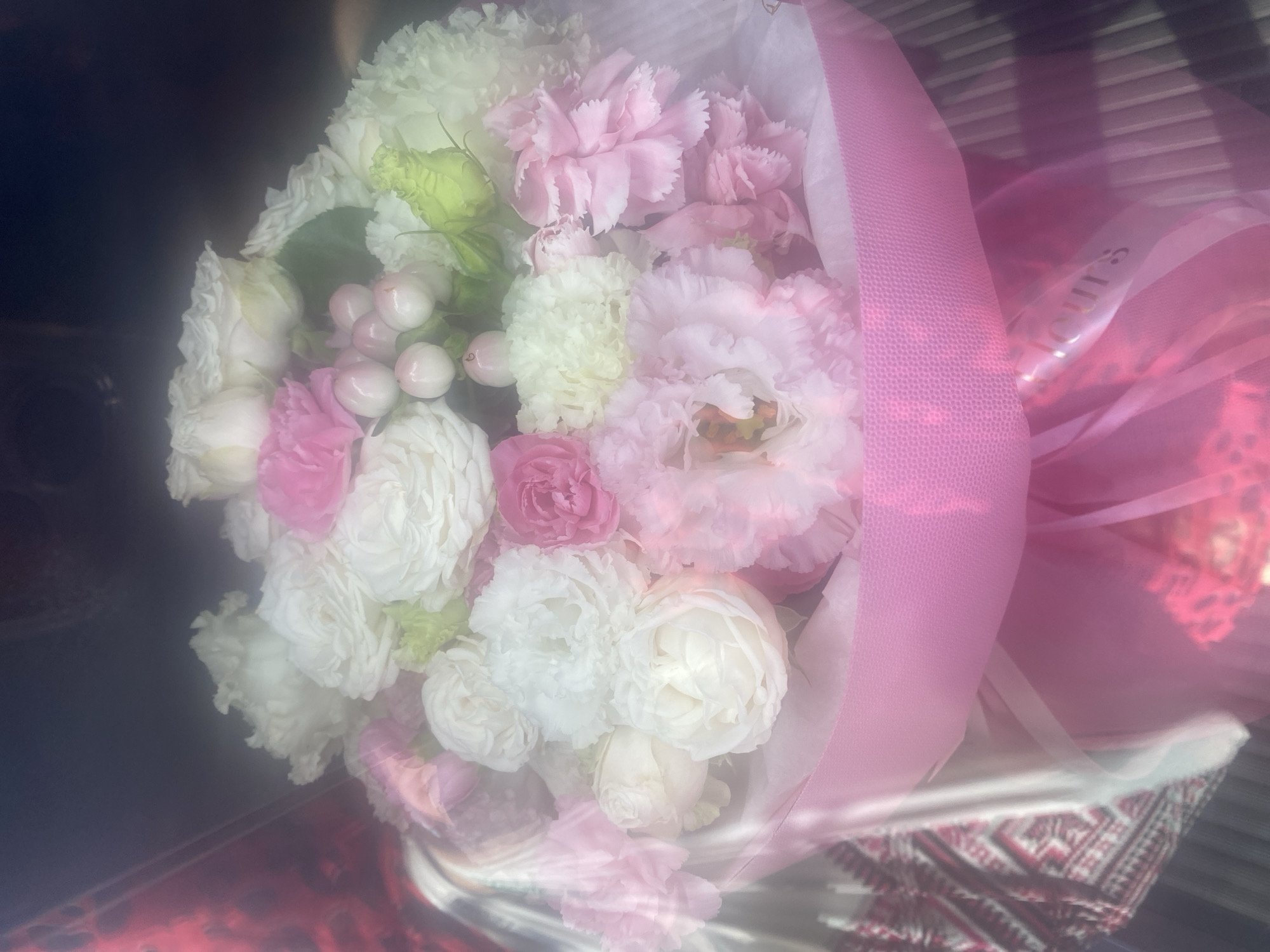A bouquet of flowers, predominantly pink and white, is presented in a clear plastic wrap tied with a pink ribbon. The bouquet contains various types of flowers, including roses, carnations, and small white berries. The background is dark and out of focus, with hints of patterned fabric visible on the left side. The lighting is soft, creating a diffused effect. The image does not provide any information about the city of Novi Petrivtsi, Ukraine.