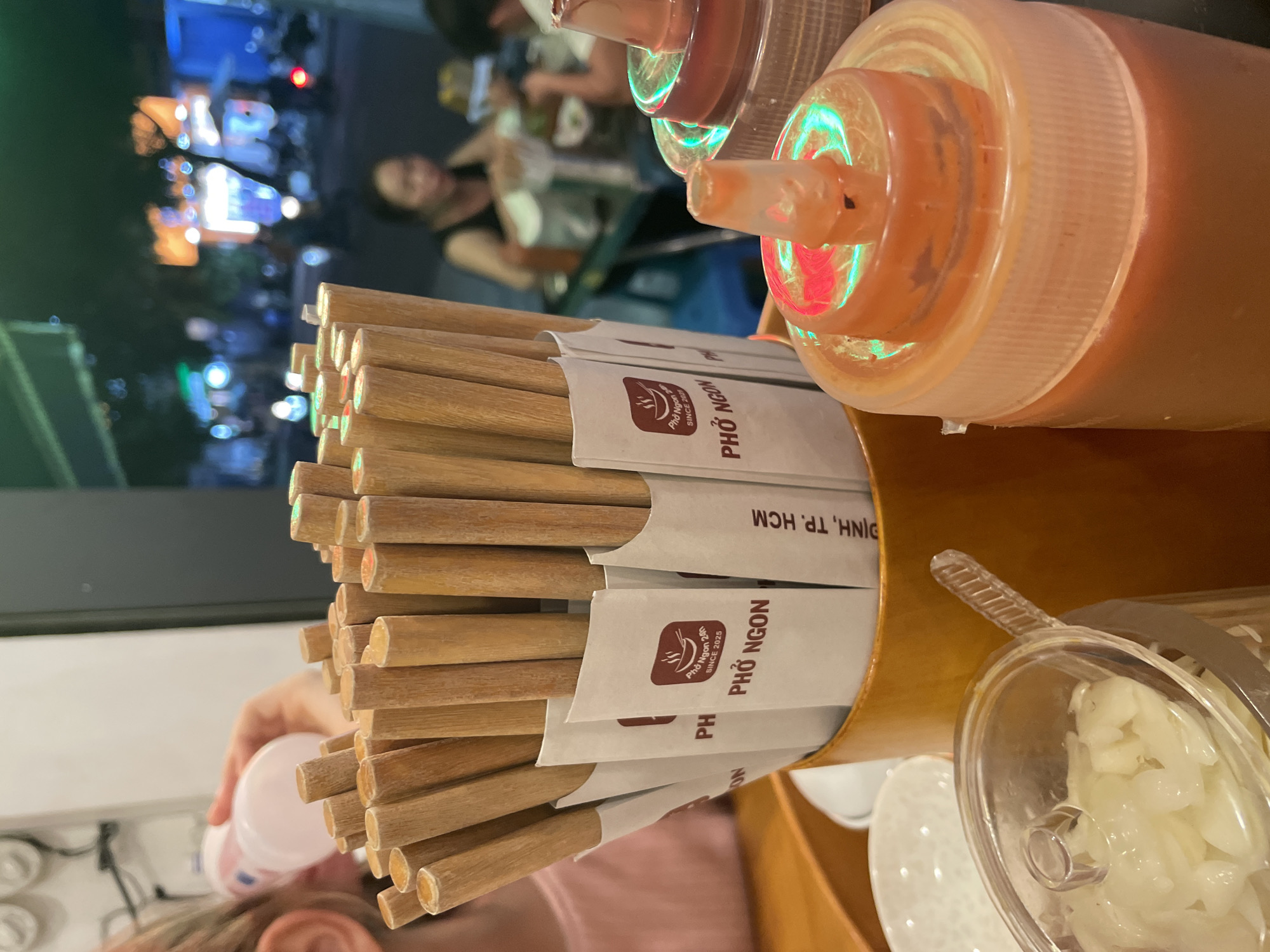 This is a close-up, eye-level, indoor shot of chopsticks in a holder on a table at a restaurant, possibly during the evening. The chopsticks are wrapped in white paper sleeves with the text "Phở Ngon 24h SINCE 2025" and a logo of a bowl with steam, alongside "PHỞ NGON" and "ĐỊNH, TP. HCM".
In the background, the scene is out of focus but suggests a busy restaurant with people dining. Lights from outside are visible, indicating it might be nighttime. To the right, there are large squeeze bottles, likely containing sauces, with one bottle cap reflecting light, showing hints of green and red. A clear container filled with chopped white ingredients, possibly garlic or onions, is also visible on the right side of the frame. A hand is seen in the lower left corner, holding a small white bottle. The overall impression is of a casual dining establishment serving Vietnamese cuisine, with a focus on pho. The "TP. HCM" indicates the location is Ho Chi Minh City, and "Thủ Đức" specifies the district within Ho Chi Minh City.